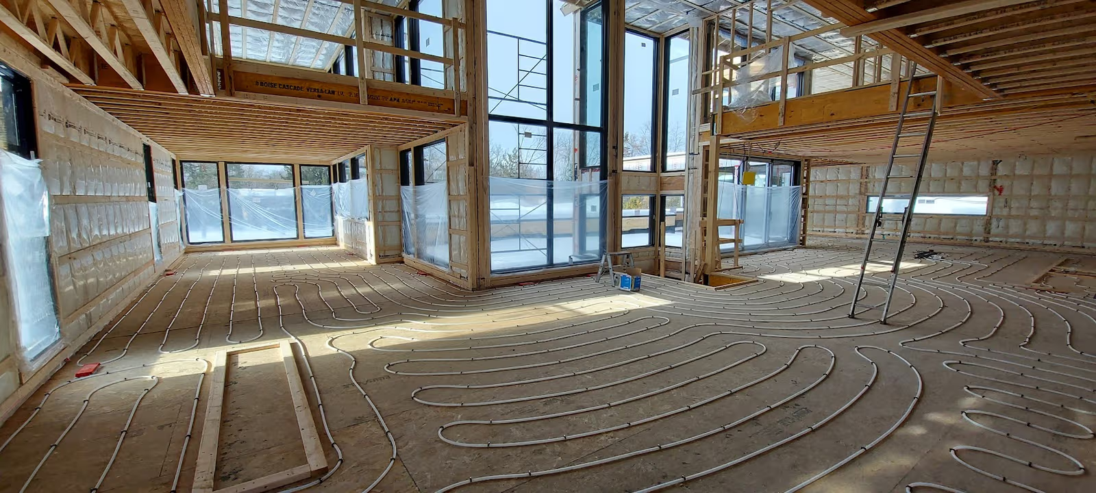 Installation de plancher chauffant hydronique dans une maison en construction avec charpente apparente, mezzanines, grandes fenêtres et tuyaux au sol