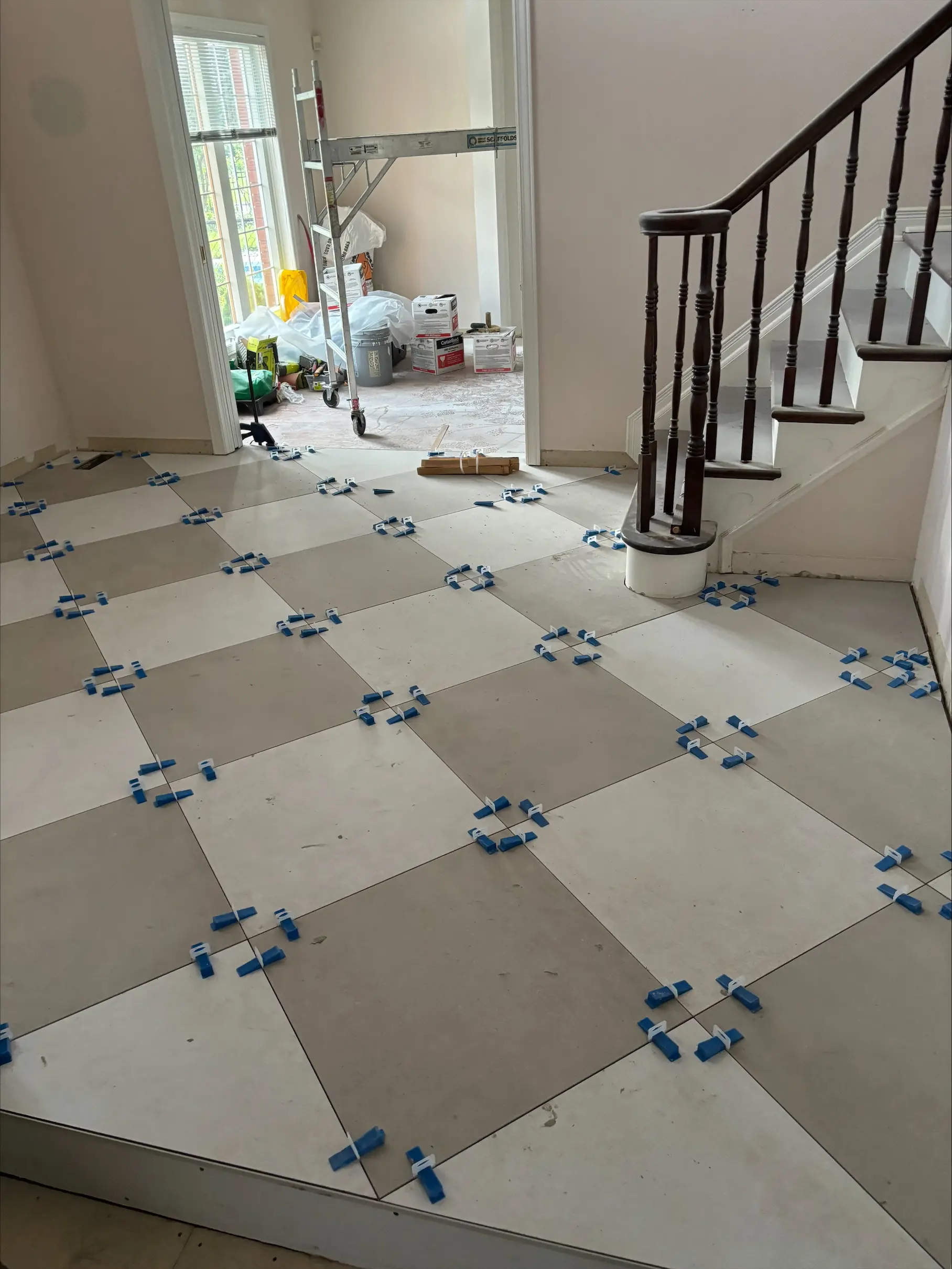 Pose de grands carreaux de céramique beige dans une entrée avec systèmes de nivellement bleus, escalier en bois foncé et matériel de rénovation en arrière-plan.
