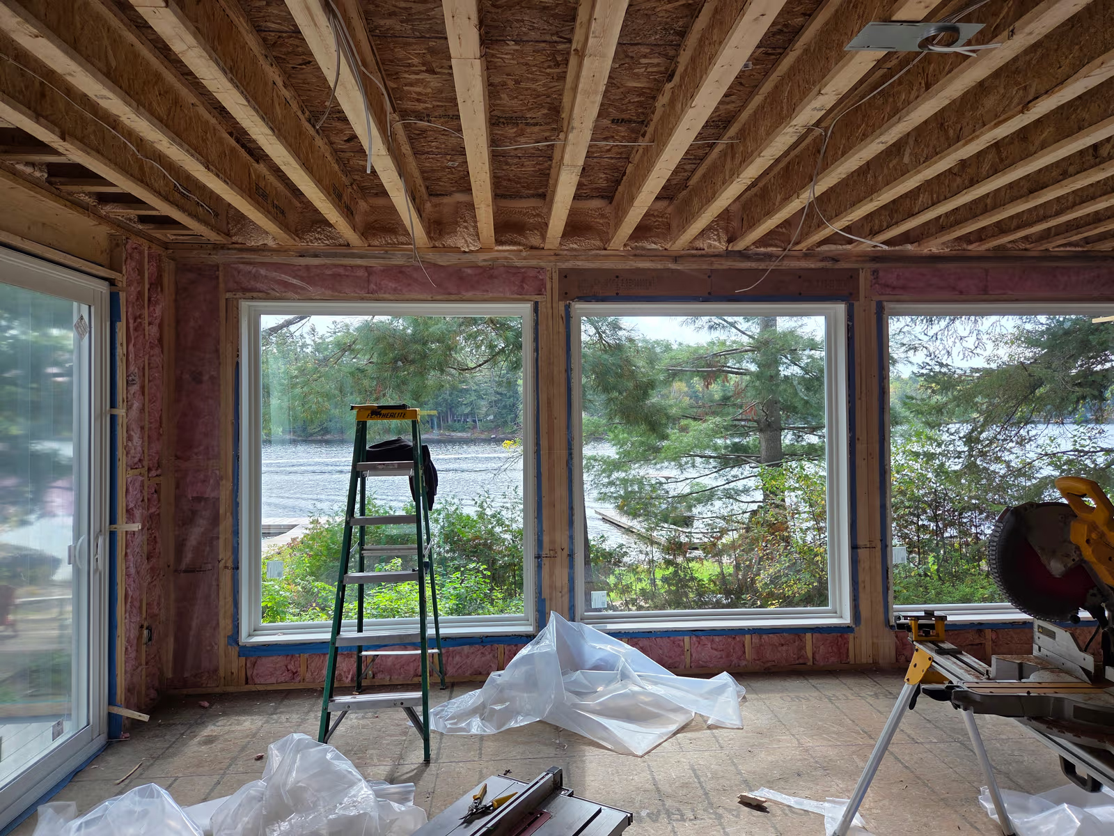 House interior under renovation with exposed ceiling framing, wall insulation, large waterfront windows, and construction tools.