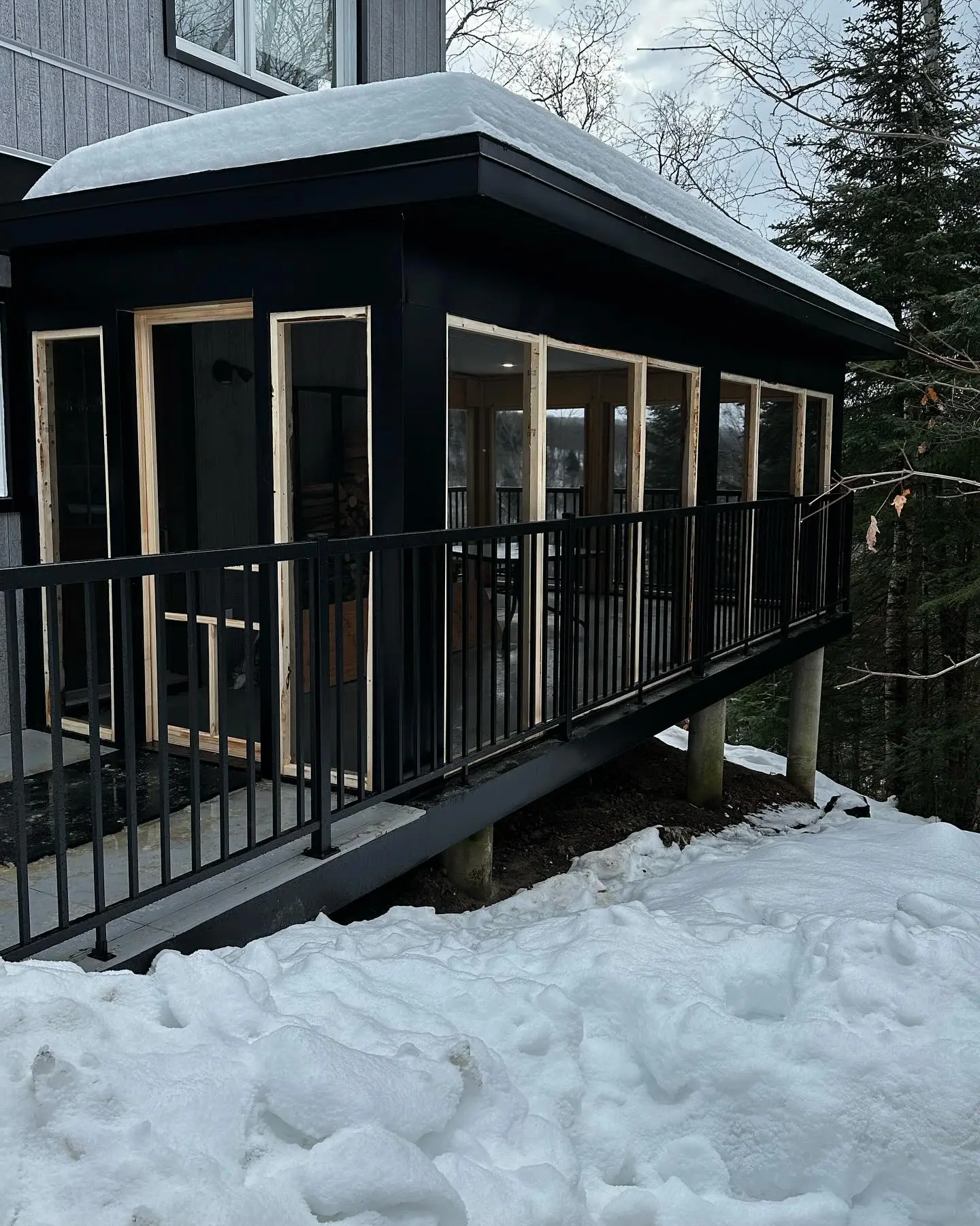Véranda moderne en construction avec structure sur pilotis et fenêtres panoramiques, entourée de neige, pour une maison contemporaine et lumineuse. 