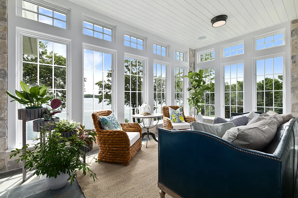 Véranda lumineuse avec grandes fenêtres panoramiques, vue sur lac et décor élégant avec mobilier en rotin et plantes vertes.