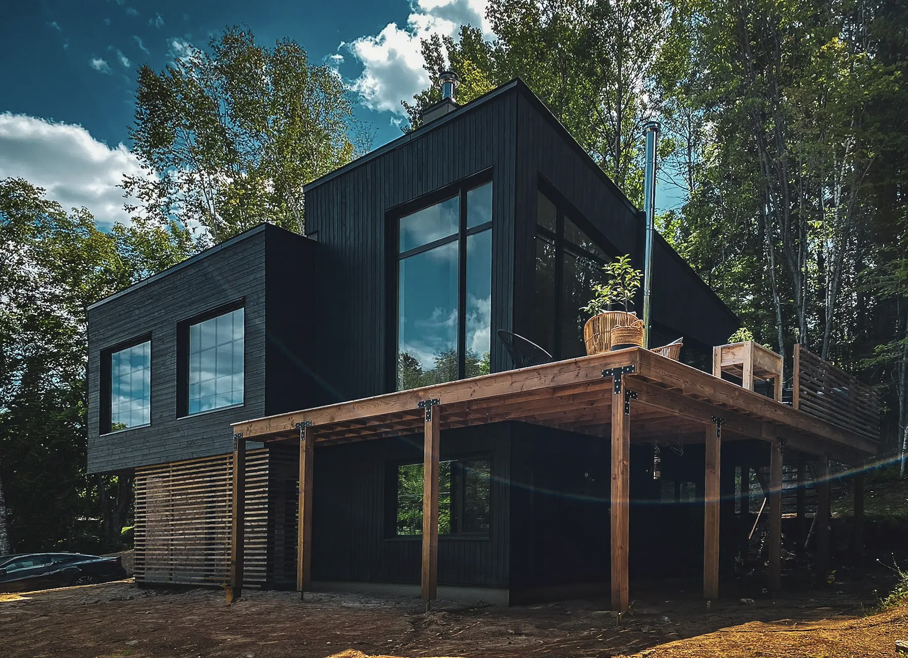 Modern house with black facade, large windows and wooden terrace on stilts, surrounded by forest