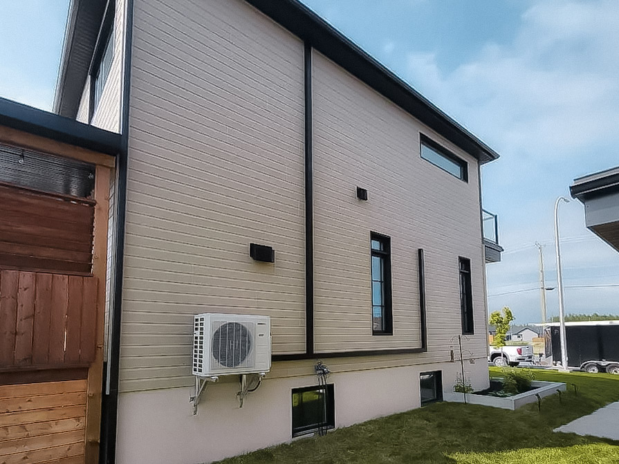 Façade latérale d’une maison moderne avec revêtement horizontal beige, fenêtres noires, unité extérieure de thermopompe et gouttières foncées.