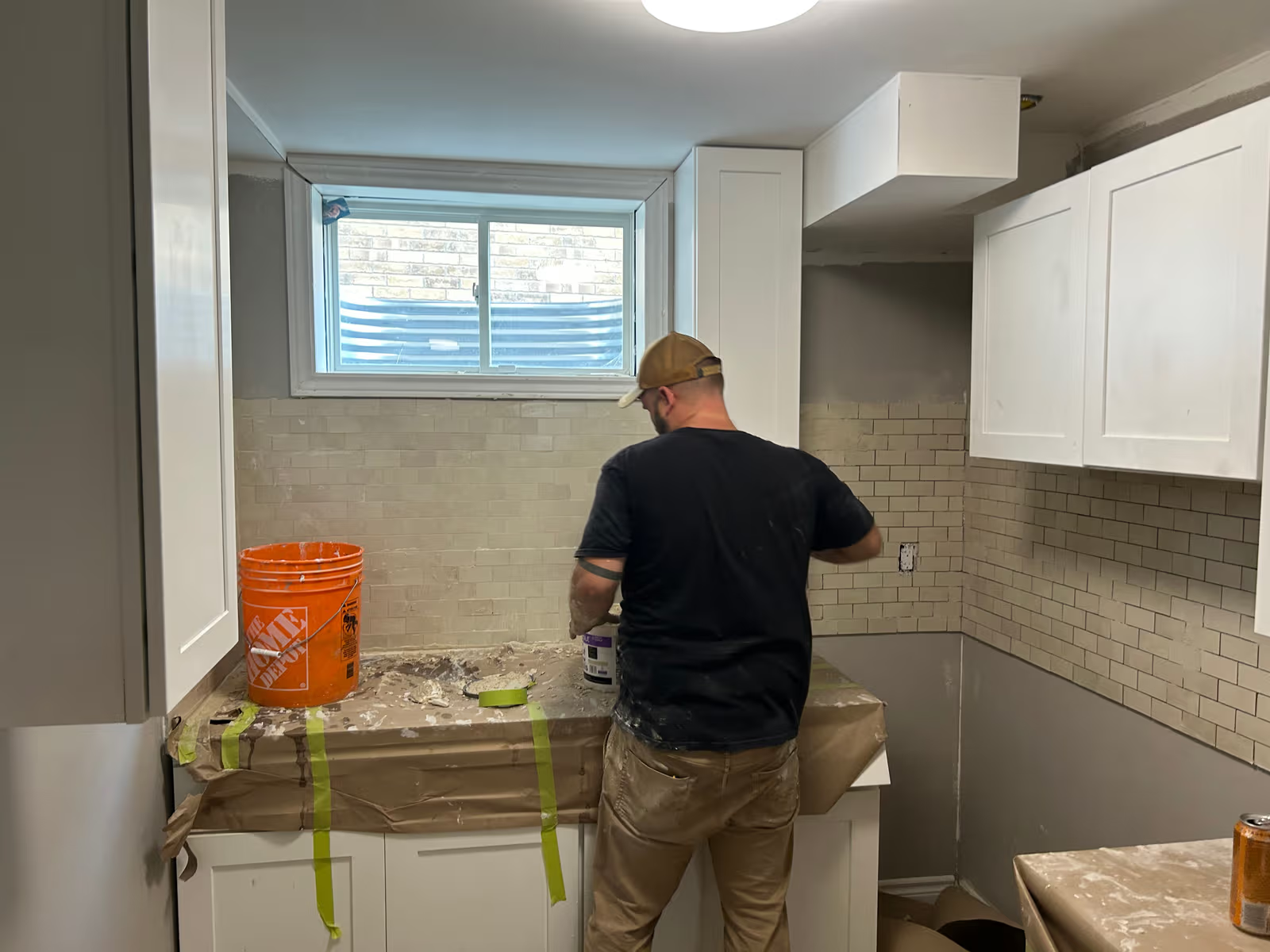 Worker applying grout to a white ceramic backsplash in a kitchen under renovation with countertops protected.