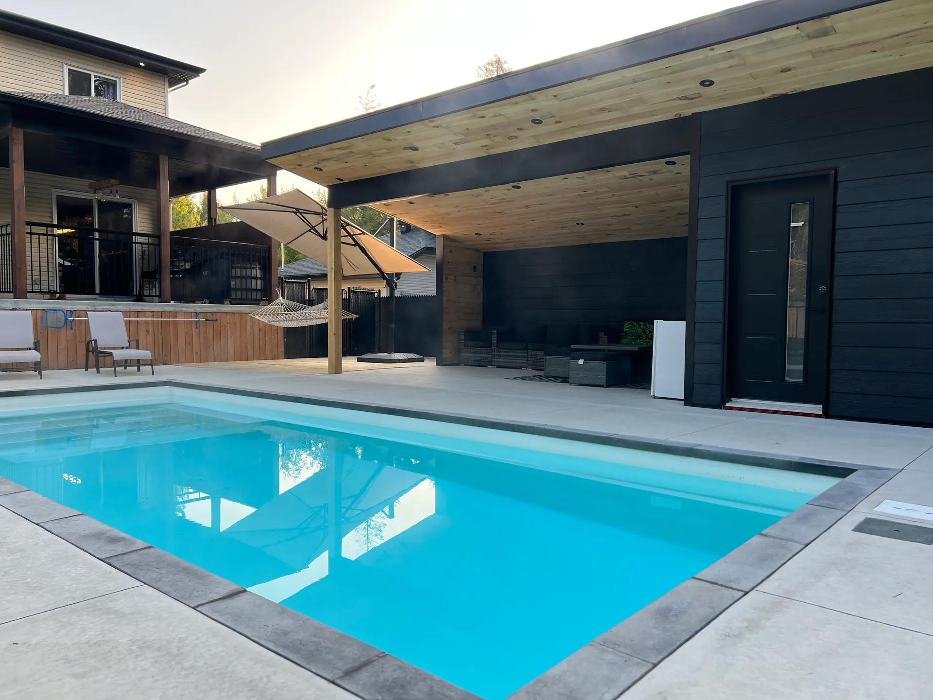 Modern backyard with inground pool, concrete slab terrace, black pavilion with wood ceiling, recessed lighting, exterior door and covered lounge area