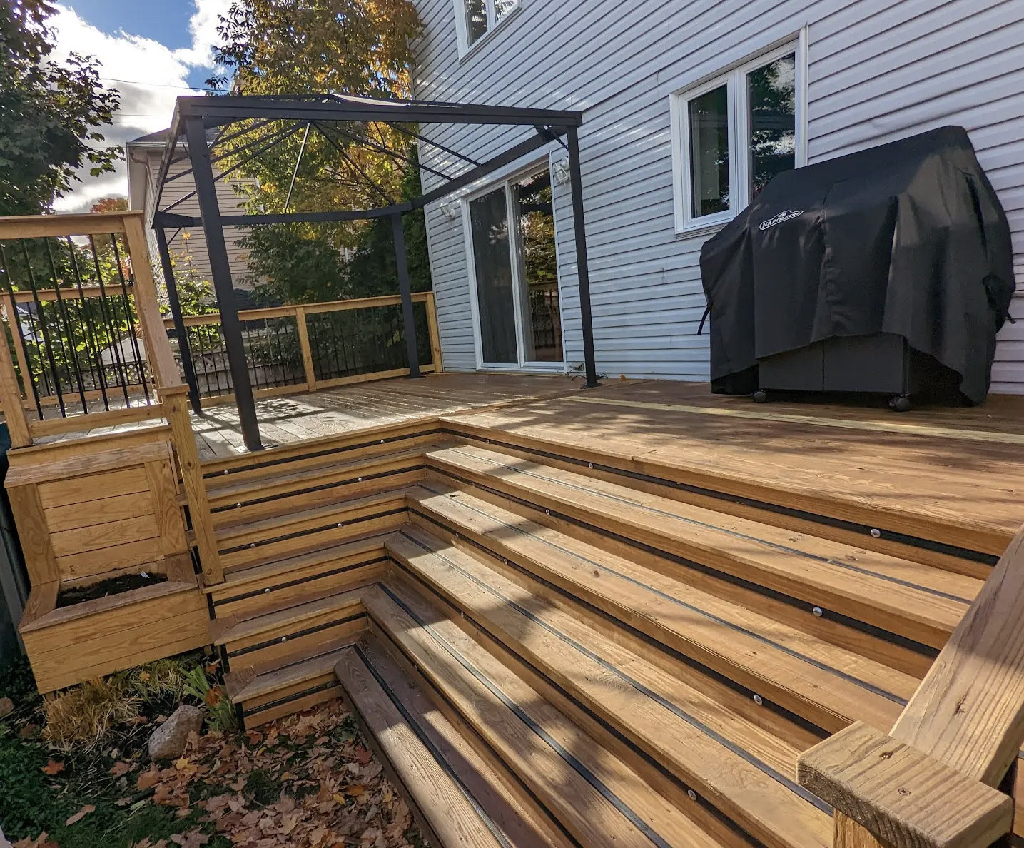Multi-level wooden outdoor deck with wide stairs, railings, pergola frame, and barbecue in a residential backyard.