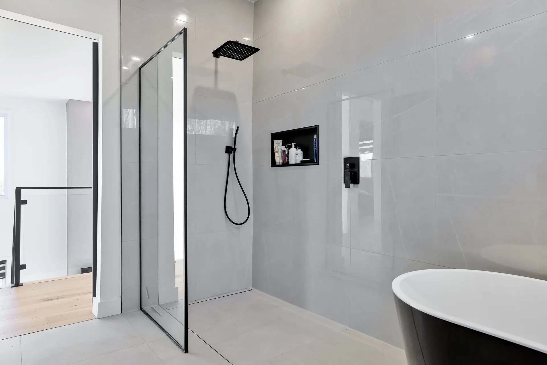Douche italienne moderne avec grandes tuiles grises, paroi de verre, robinetterie noire, niche murale intégrée et baignoire autoportante noire.