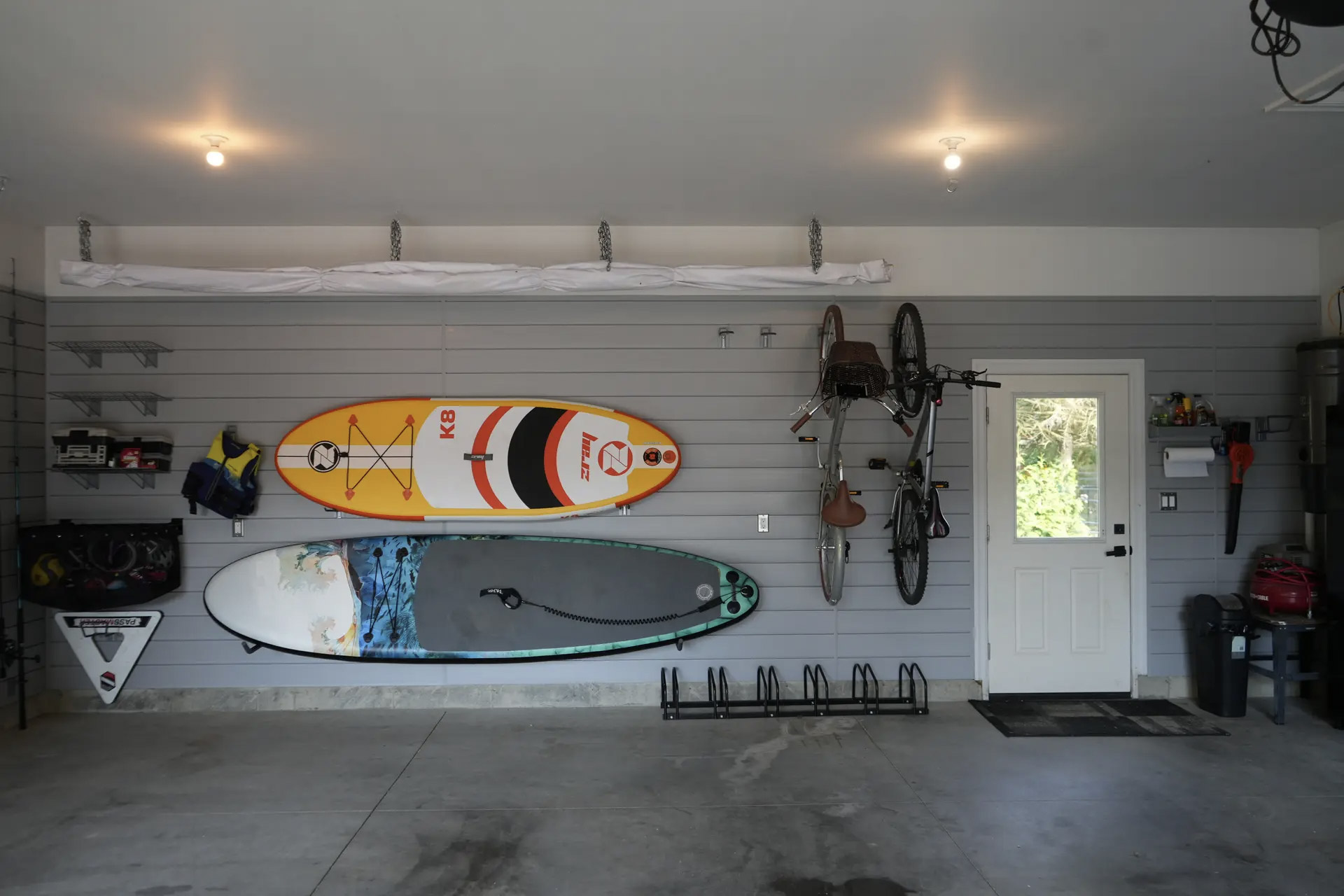 Intérieur de garage aménagé avec panneaux muraux gris, planches de surf et de pagaie, vélos suspendus, supports de rangement et porte extérieure blanche.