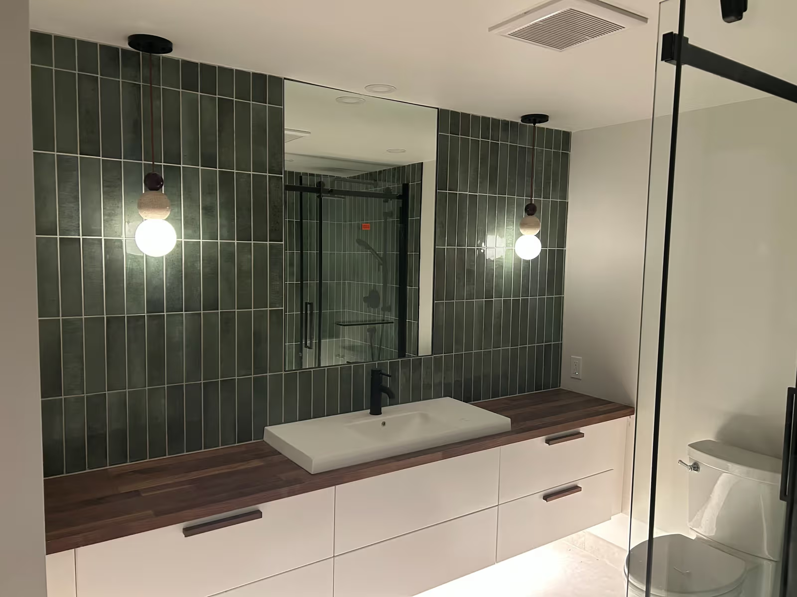 Salle de bain moderne avec meuble-lavabo blanc, comptoir en bois foncé, carrelage mural vert vertical, grand miroir, robinet noir et douche vitrée