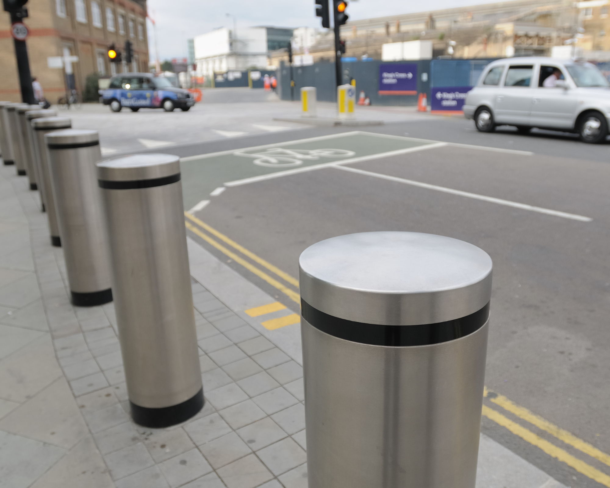 Row of stainless steel security bollards along a city sidewalk, with marked bike lane and car traffic in the background.
