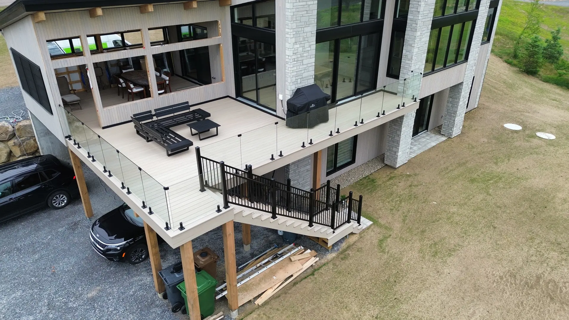 Large elevated composite deck with glass railings, stairs with black metal railings, outdoor furniture, and a contemporary façade with large windows