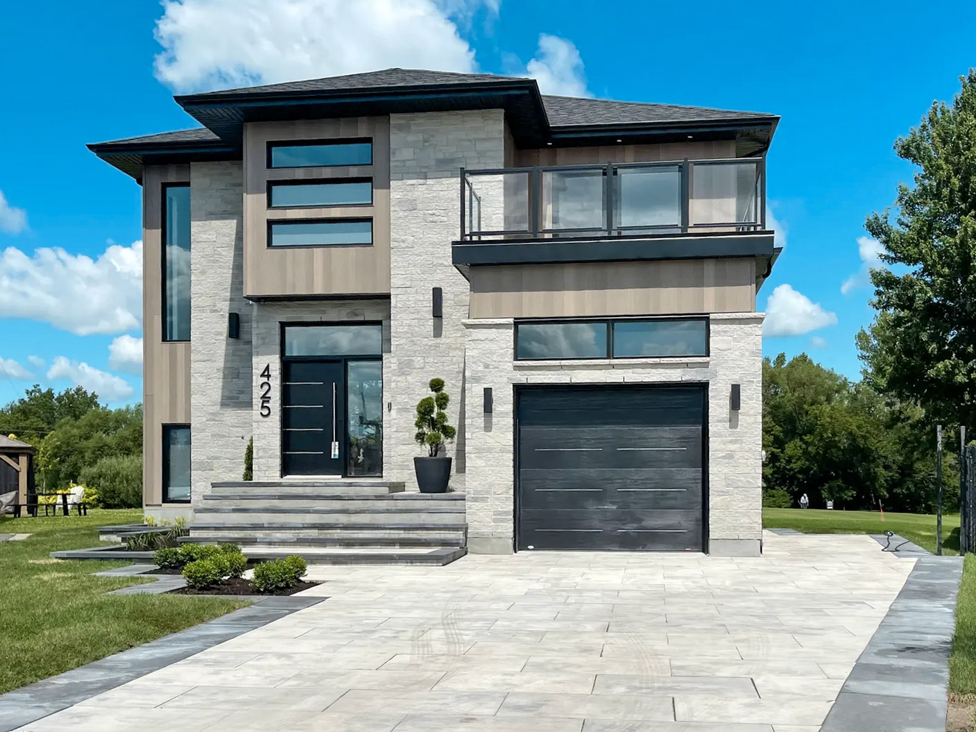 Maison contemporaine à deux étages avec façade en pierre grise, garage intégré et grande allée pavée sous ciel bleu ensoleillé