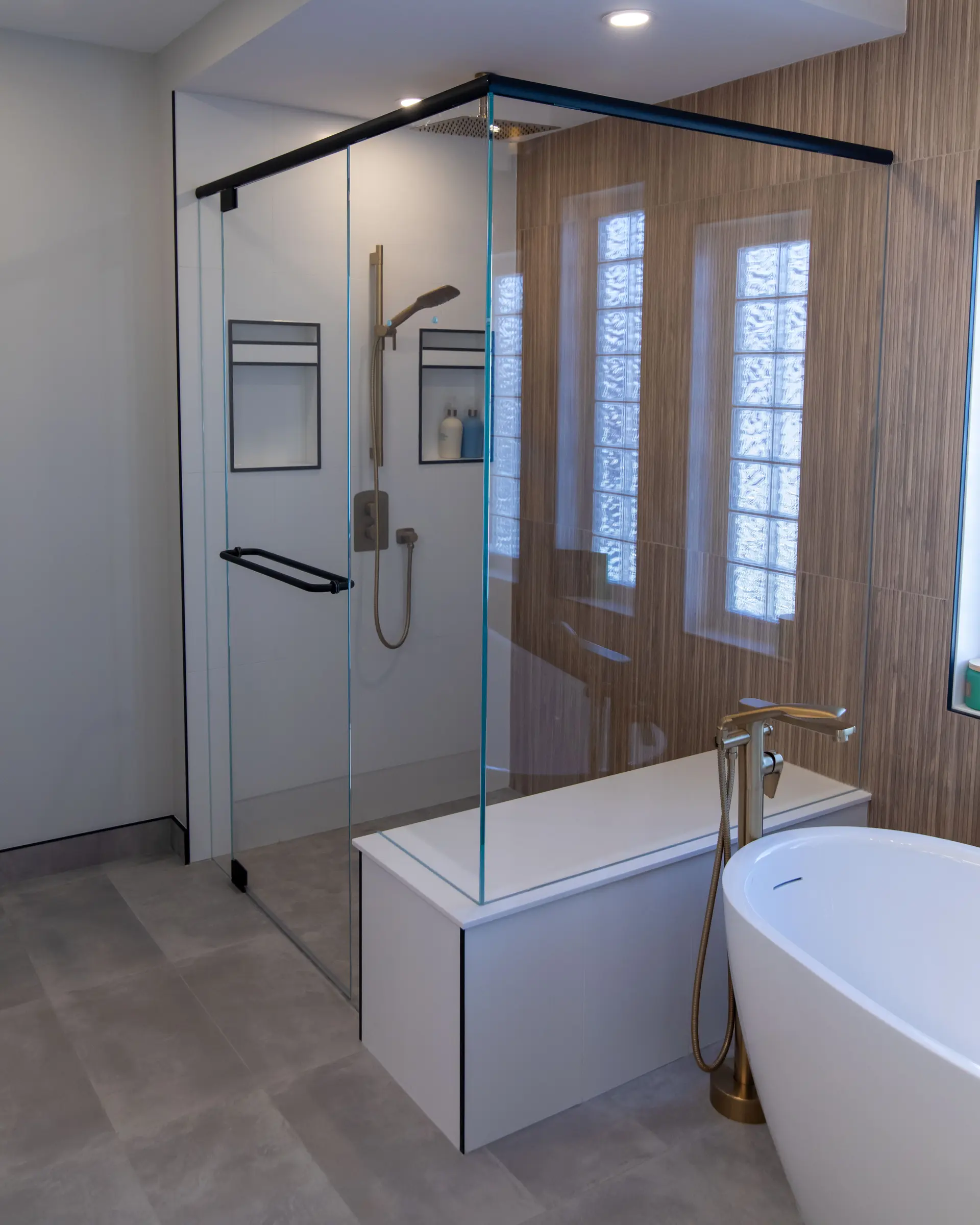 Modern bathroom with a glass shower, black hardware, wood-effect wall tiles, built-in niche, and white freestanding bathtub.