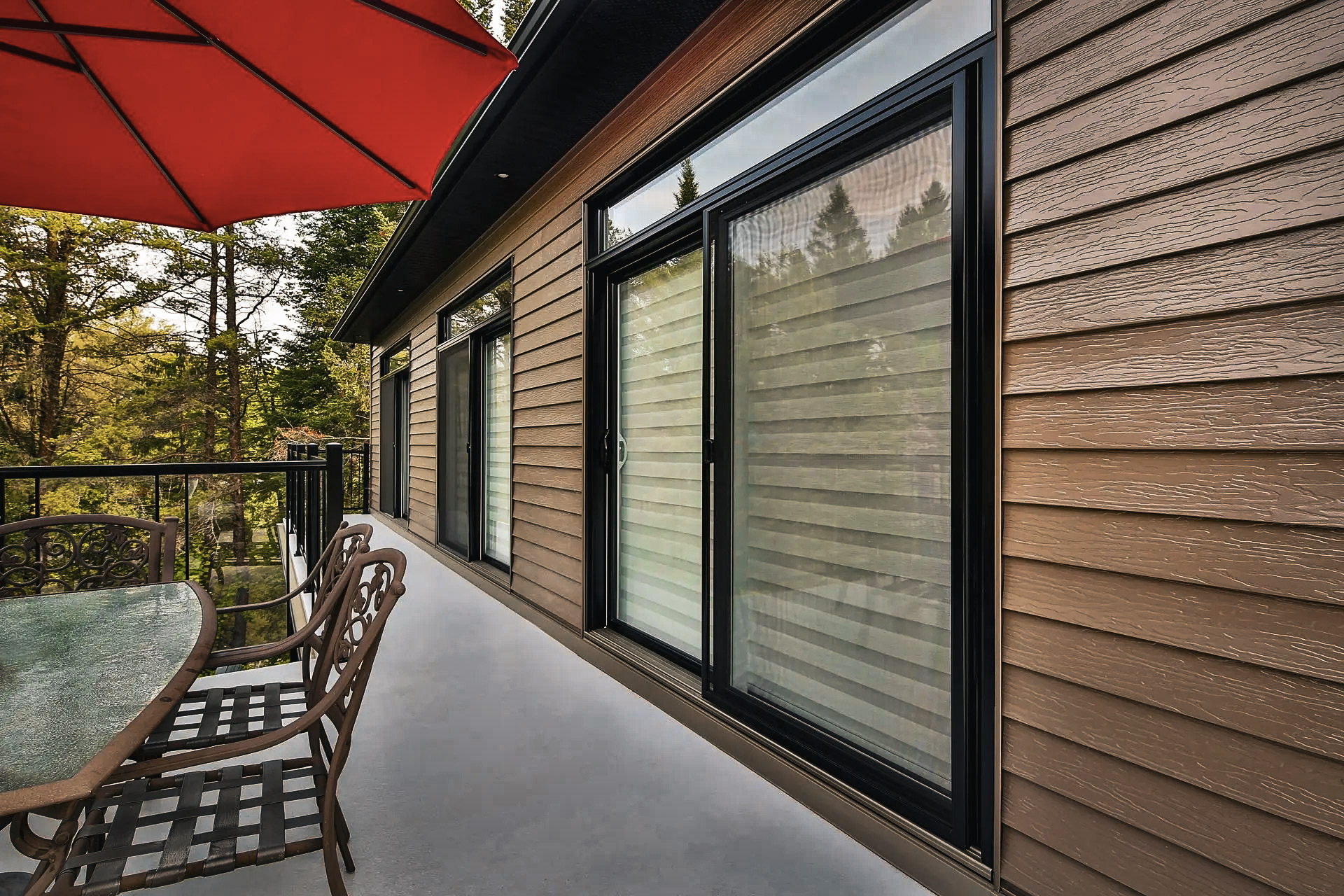 Balcon extérieur avec revêtement brun, portes patio vitrées noires, mobilier de terrasse, parasol rouge et vue sur les arbres.