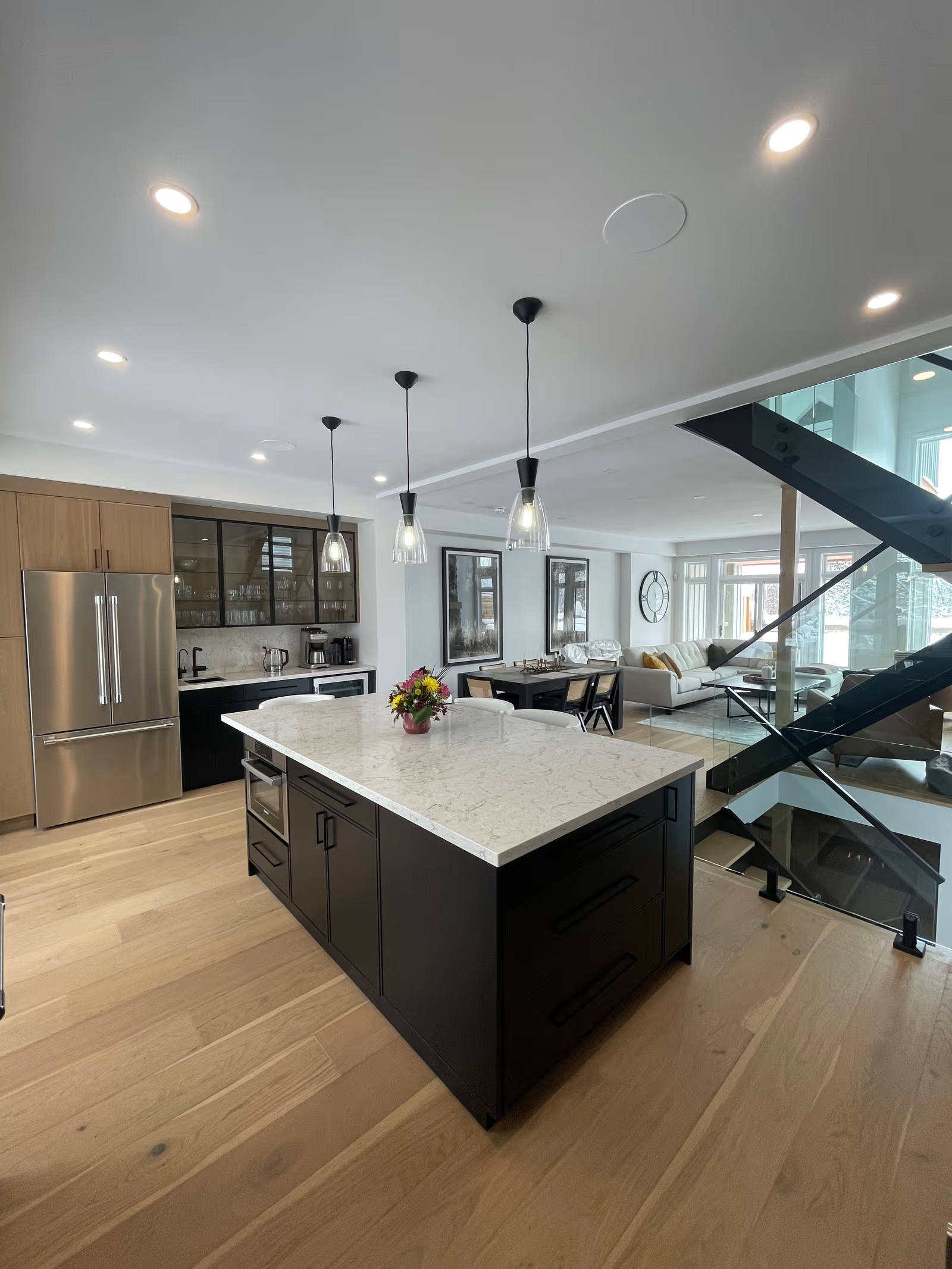 Modern open kitchen with black island, white veined countertop, glass pendant lights, wood cabinets, stainless steel appliances, and staircase with glass railing