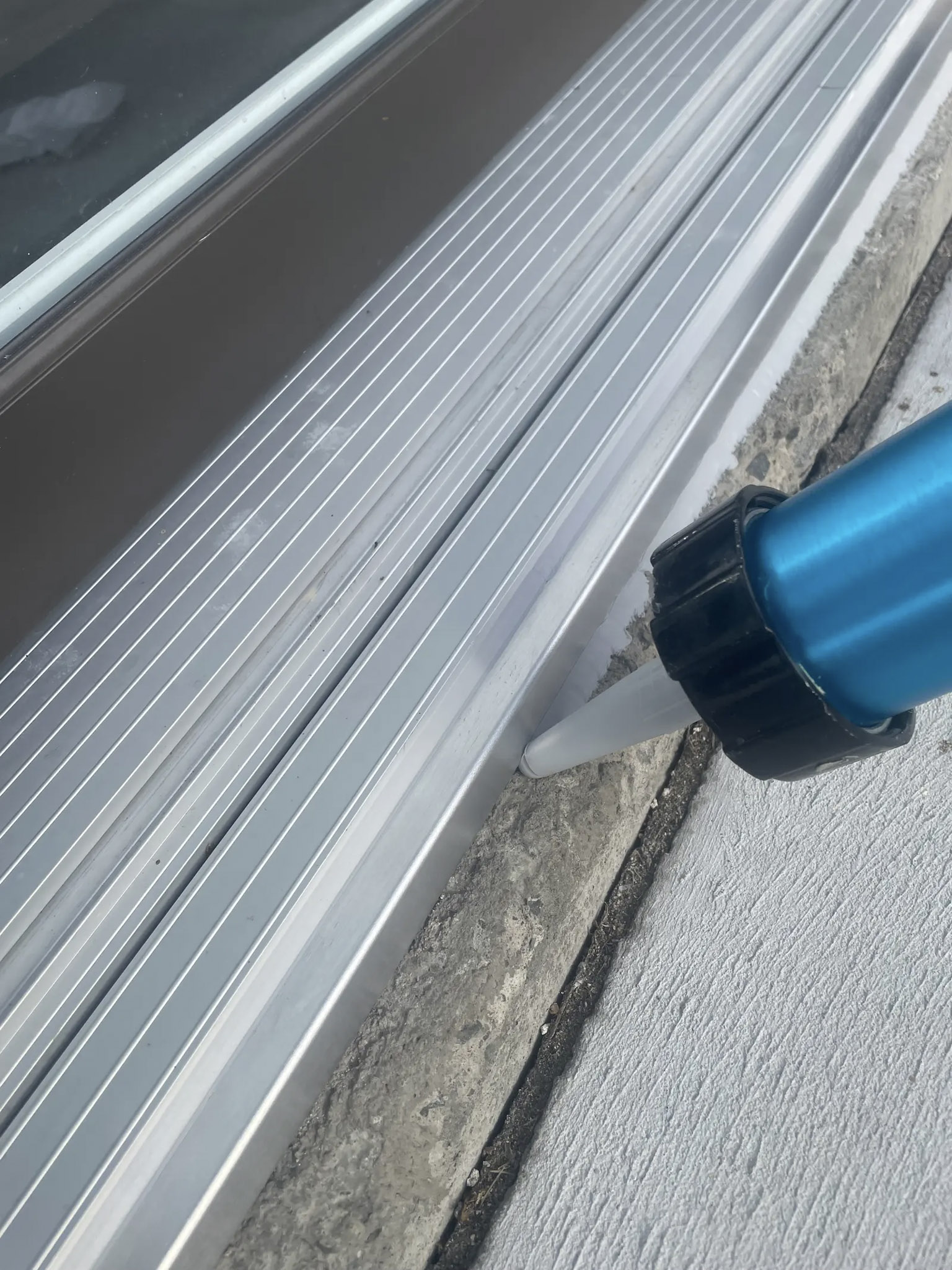 Application de calfeutrage au silicone sur le seuil en aluminium d'une porte de garage afin d'assurer l'étanchéité entre le béton et le cadre.