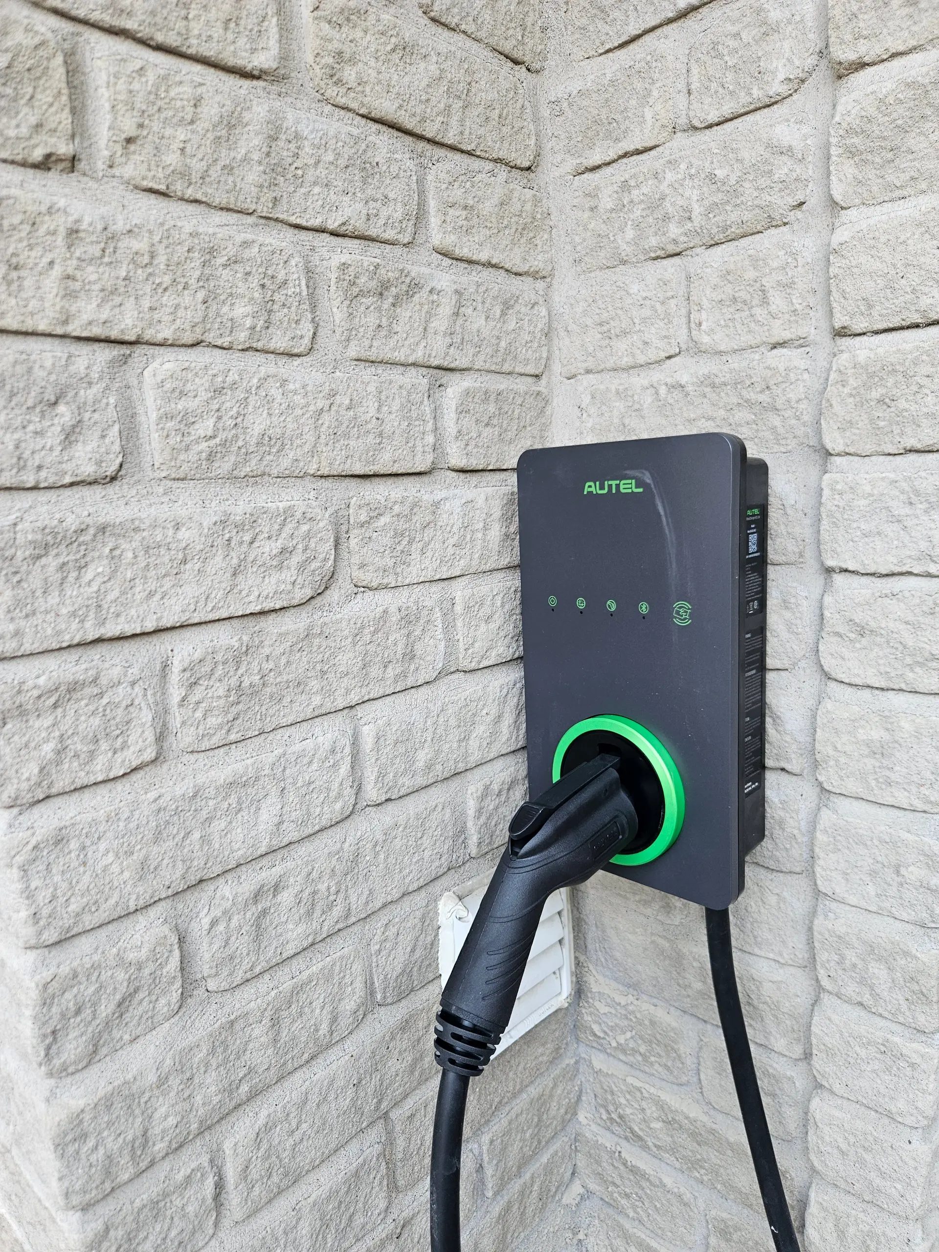 Borne de recharge pour véhicule électrique installée sur un mur extérieur en brique pâle, avec câble de recharge branché.