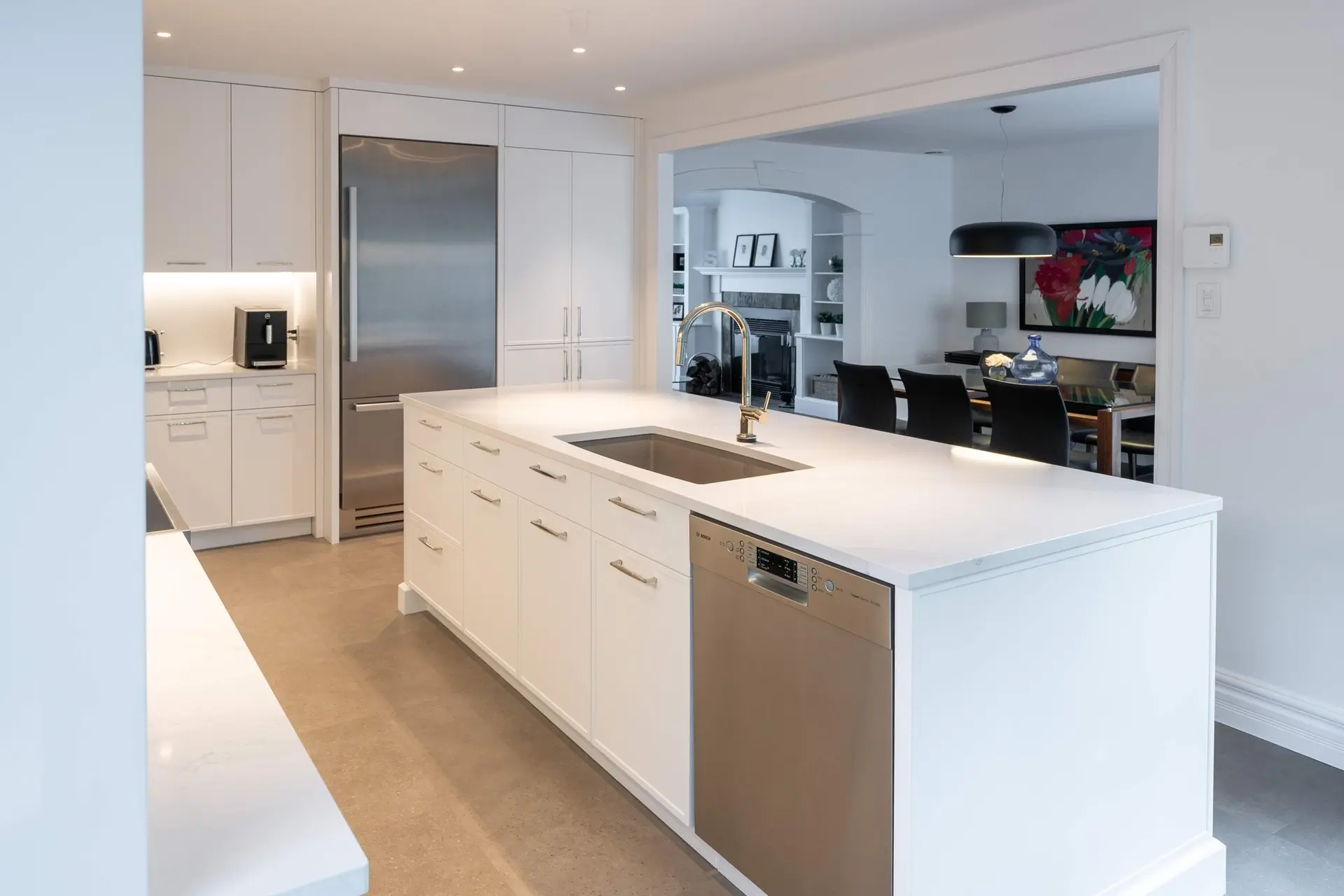 Contemporary white kitchen with large central island, light countertop, integrated sink, gold faucet, stainless steel dishwasher, and open dining room