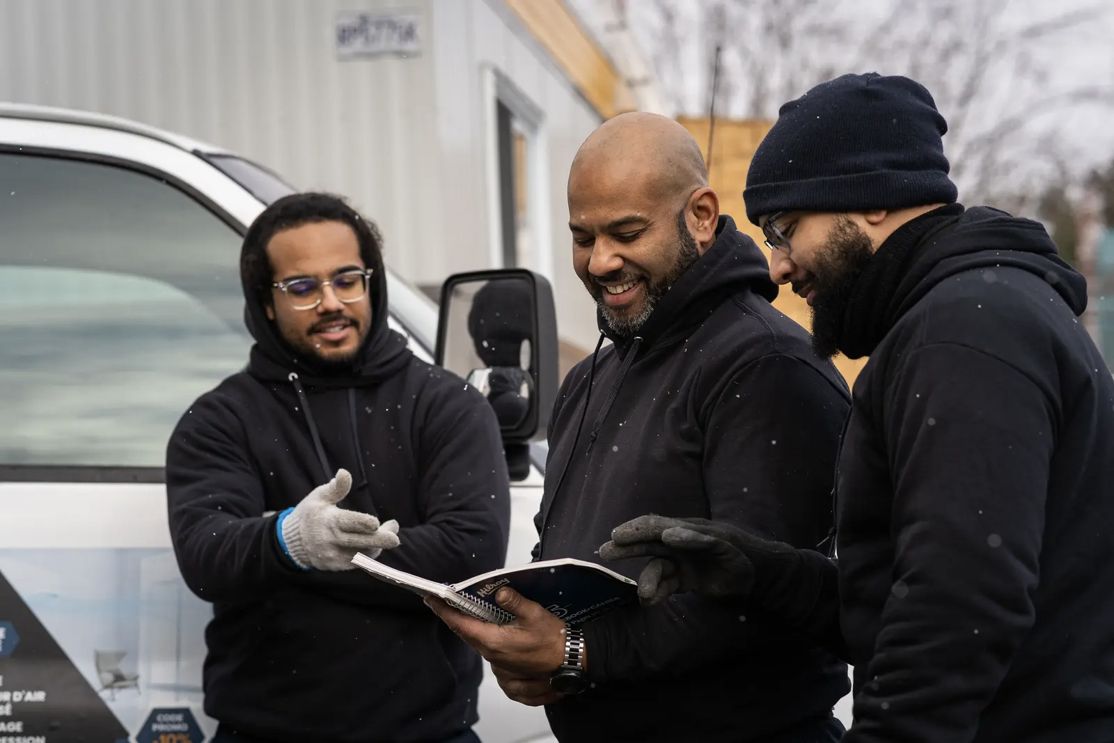 Trois ouvriers en tenue hivernale consultant un carnet devant un véhicule de chantier sur un site de construction