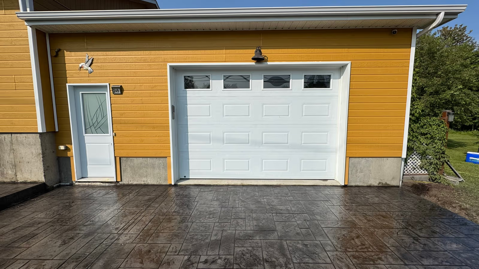 Façade de garage avec revêtement extérieur jaune, porte de garage blanche, porte d’entrée latérale et entrée en béton estampé.