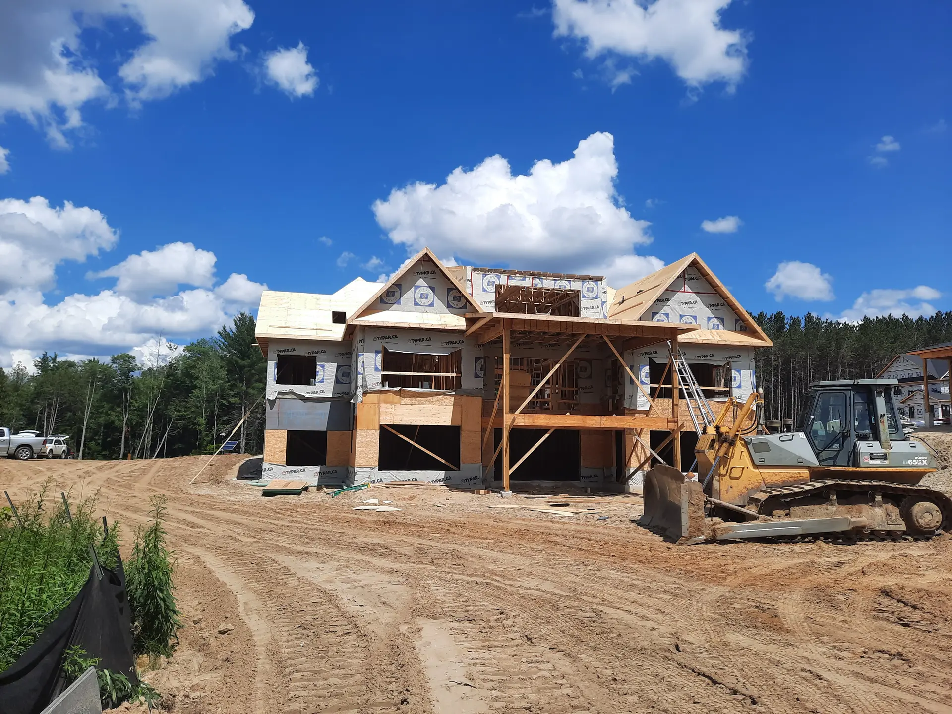 Two-story house under construction on a sandy lot with Tyvek weather wrap, wooden scaffolding, and heavy machinery on site.