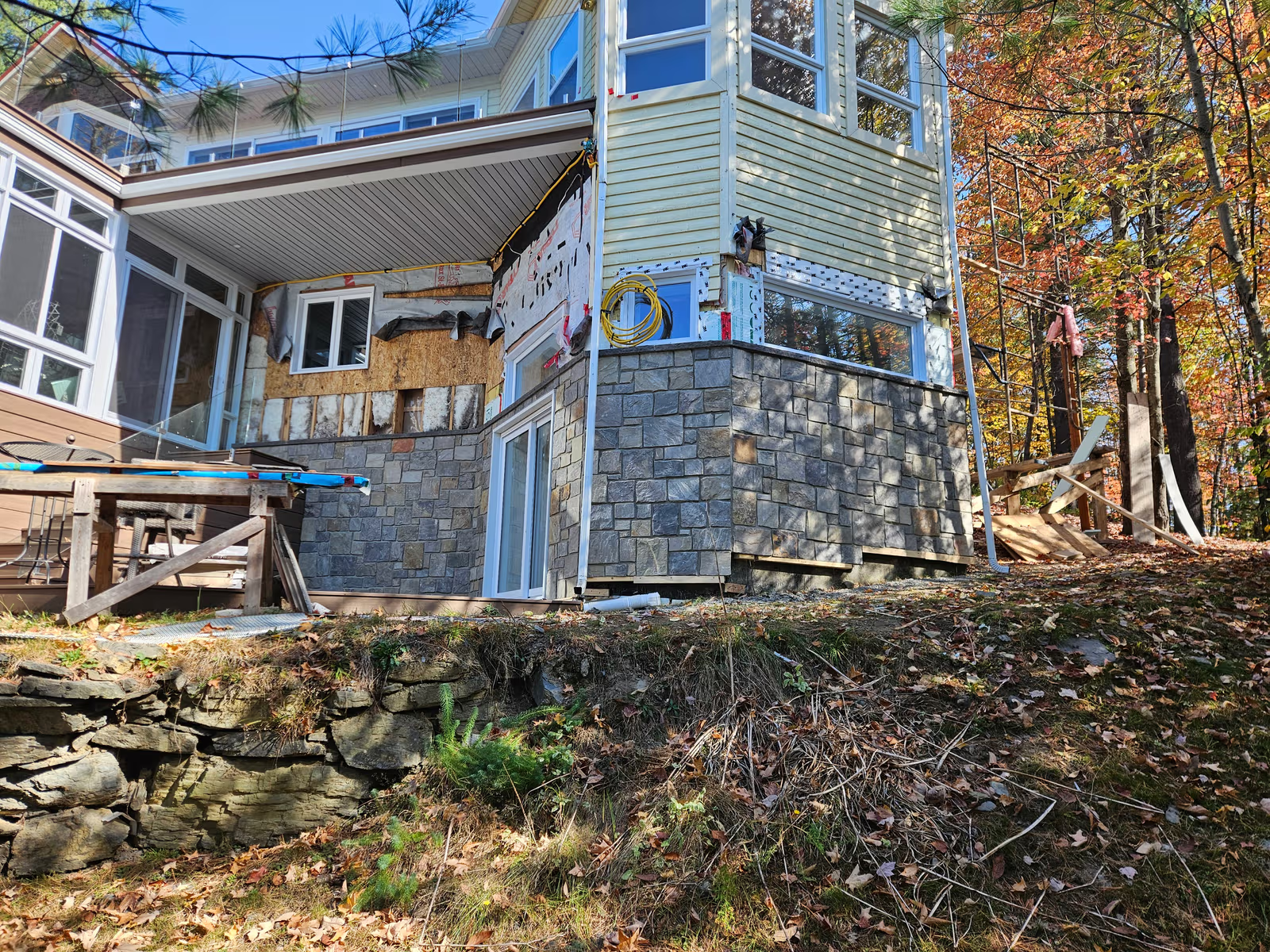 House undergoing exterior renovation with a multi-level extension, partially installed stone veneer, exposed weatherproofing membrane, and scaffolding beside a wooded lot.