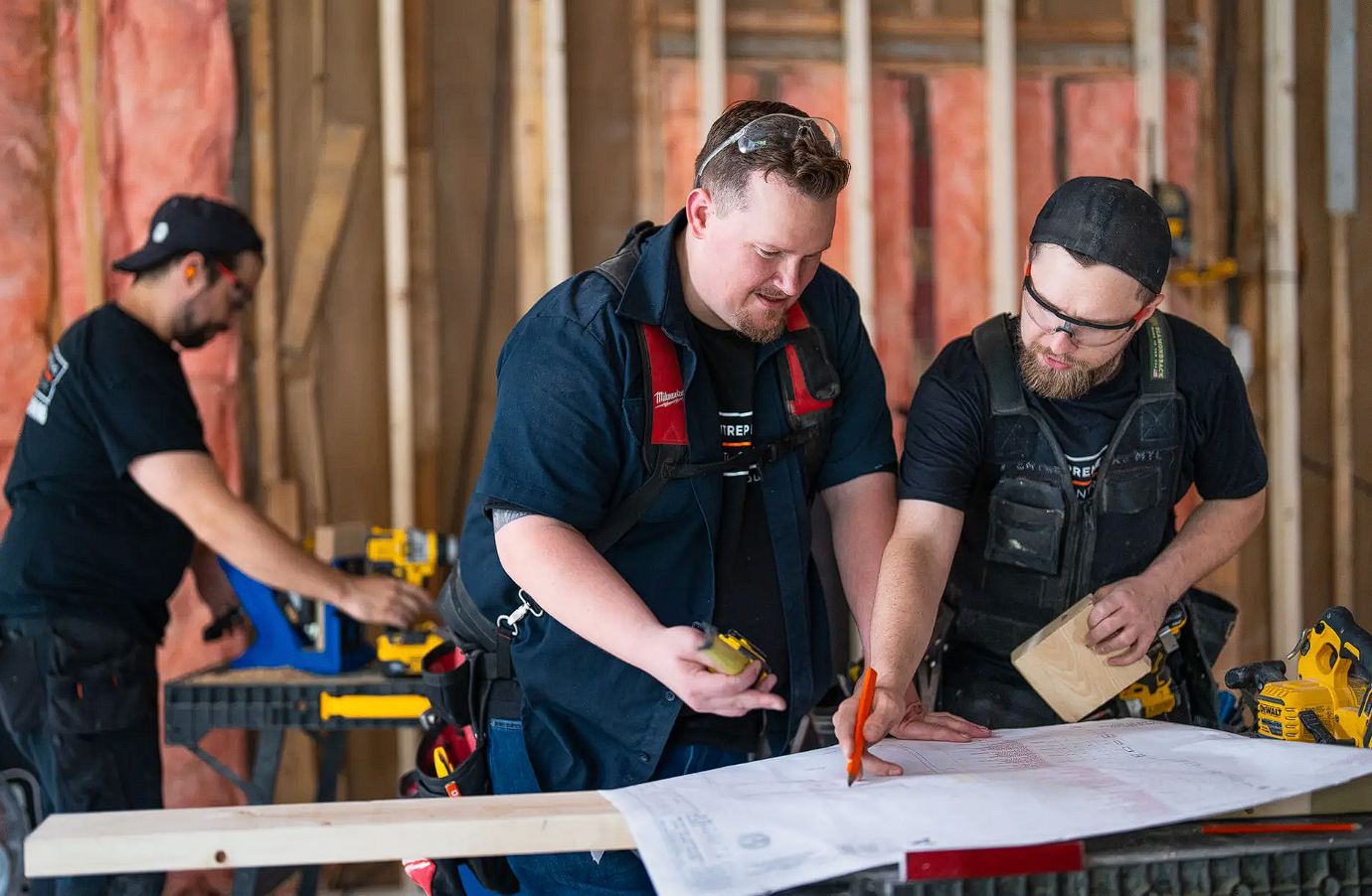 Trois ouvriers de construction analysant des plans de rénovation intérieure sur un chantier en cours avec outils et matériaux visibles