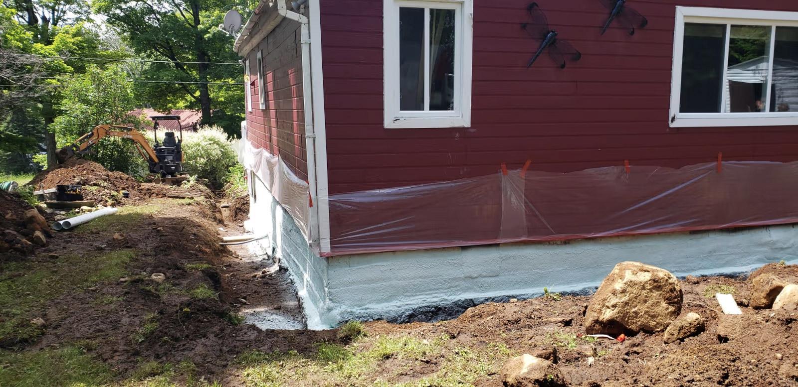 Excavation and waterproofing work around a house foundation, with a mini excavator, perimeter trench, and protective coating applied to the concrete.