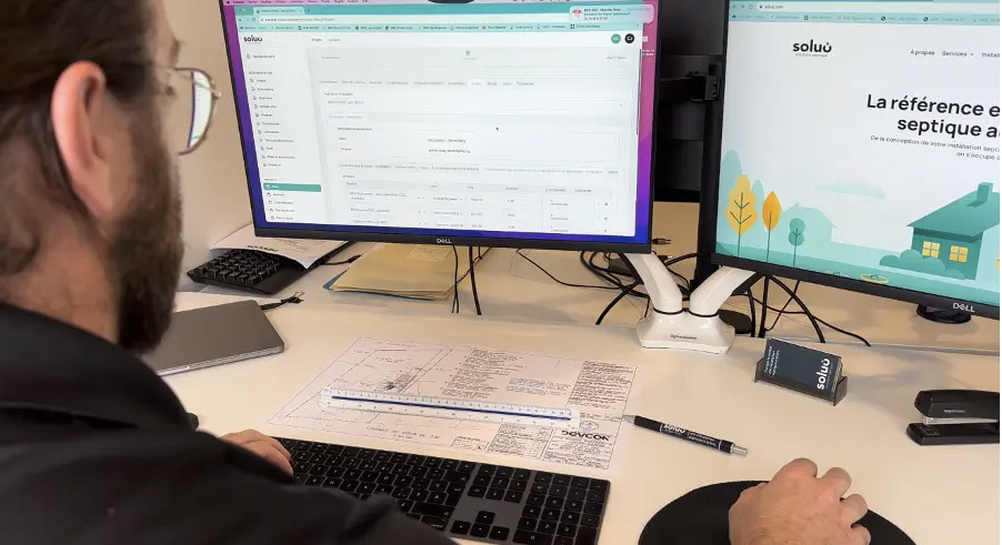 Homme travaillant à un poste informatique avec deux écrans affichant un logiciel de gestion et un site de services septiques, avec un plan technique posé sur le bureau.