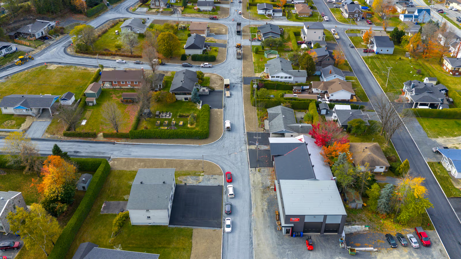 Vue aérienne d’un quartier résidentiel avec maisons, rues asphaltées, arbres colorés, terrains verts et bâtiment commercial