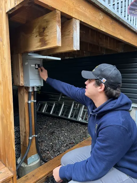 Électricien vérifiant un panneau électrique extérieur sous une terrasse en bois lors d’une installation résidentielle.