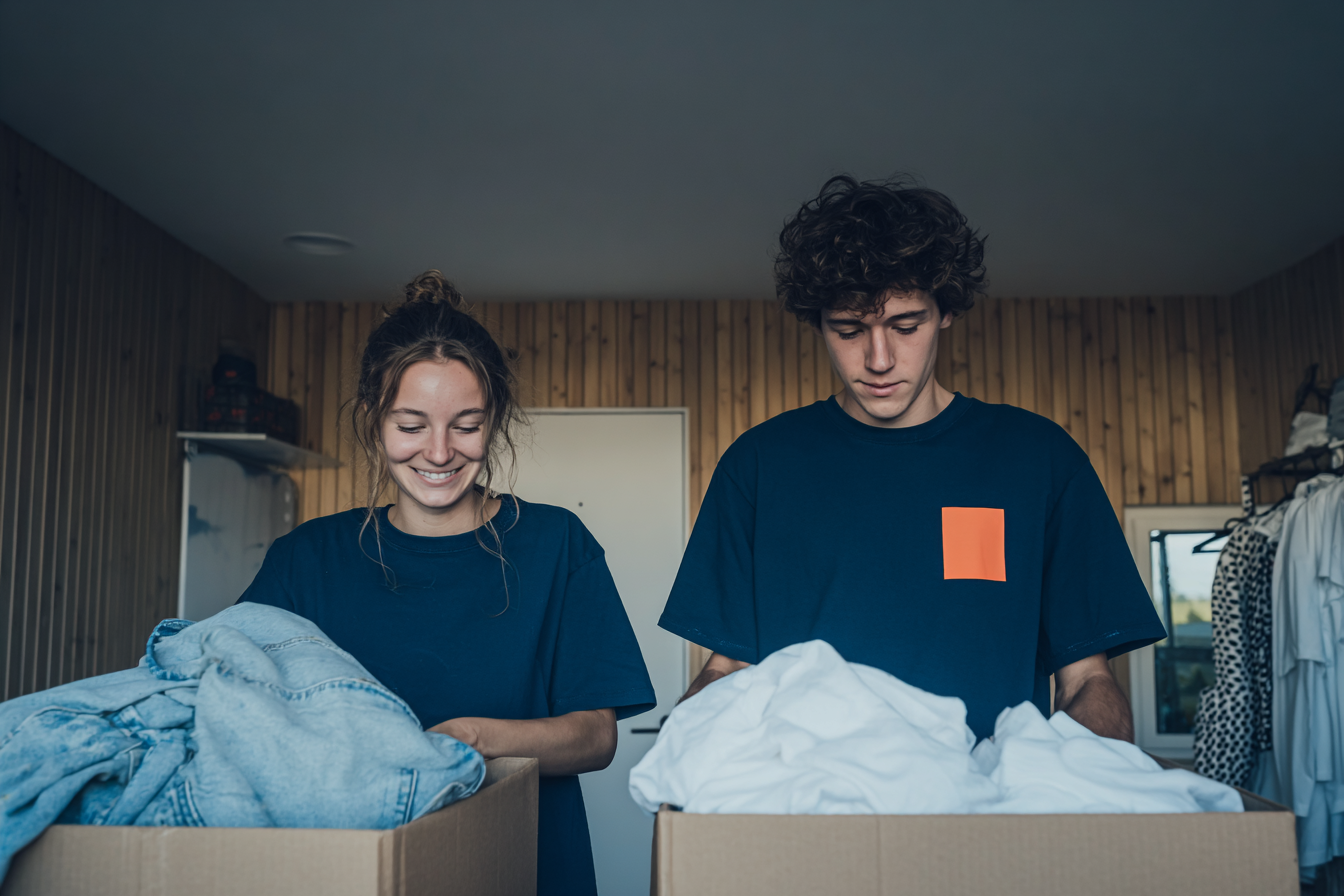 Two young people sorting clothes into cardboard boxes in a wood-paneled room
