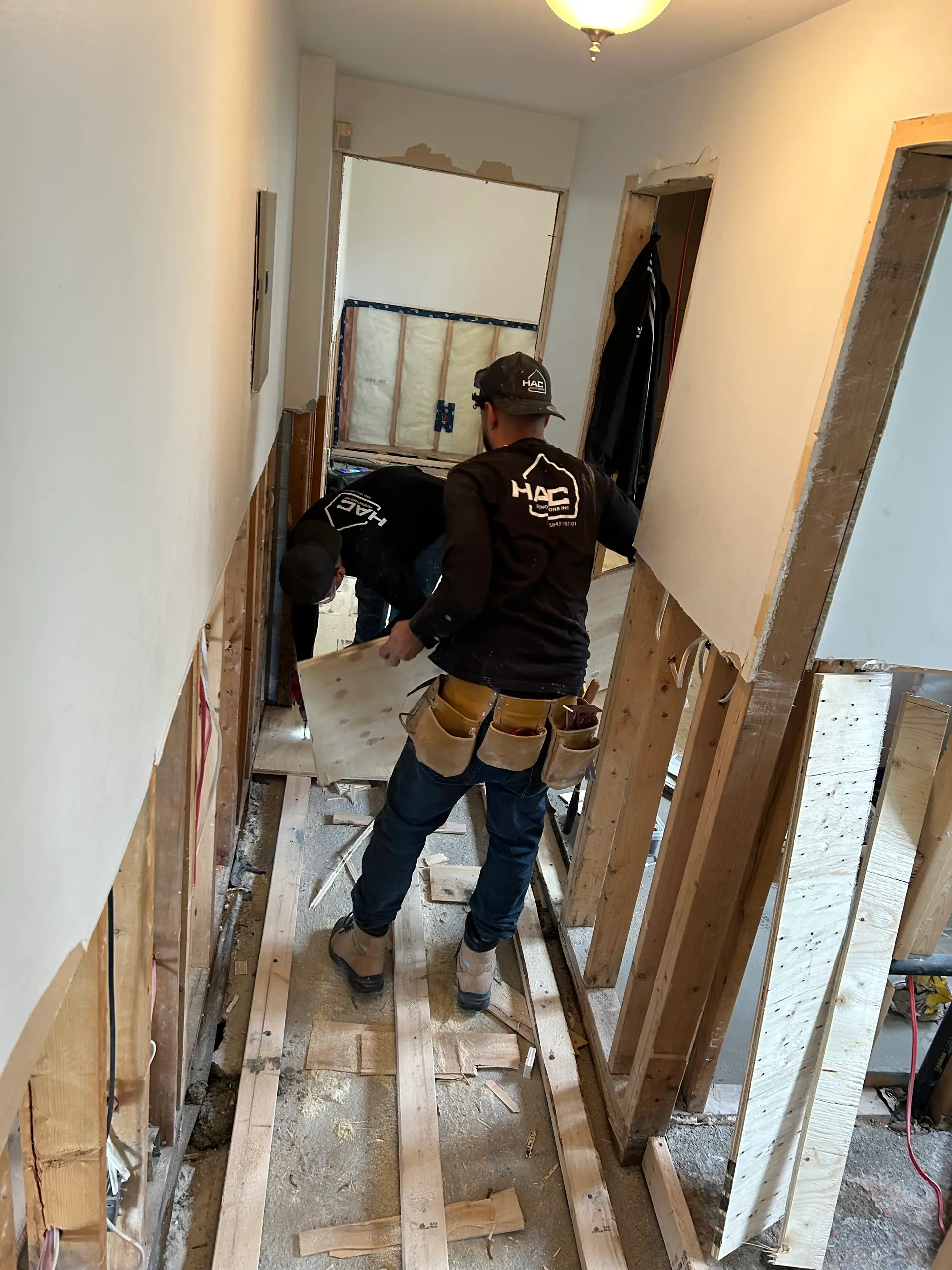 Workers renovating an interior hallway after demolition, with open walls, exposed wood framing, and construction materials on the floor.