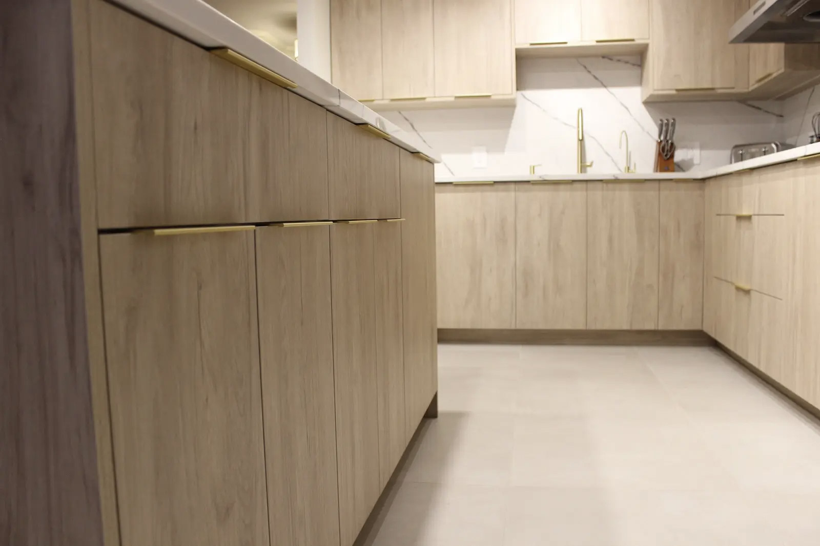 Cuisine moderne en bois clair avec armoires sans poignées apparentes, accents dorés, comptoirs blancs et dosseret effet marbre.