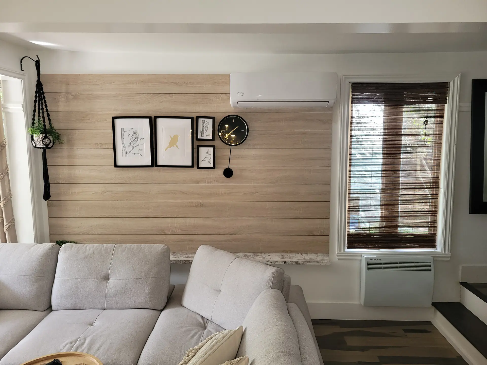 Light wood wall with wall-mounted air conditioner, decorative frame gallery, black designer clock, and hanging plant in a modern living room.