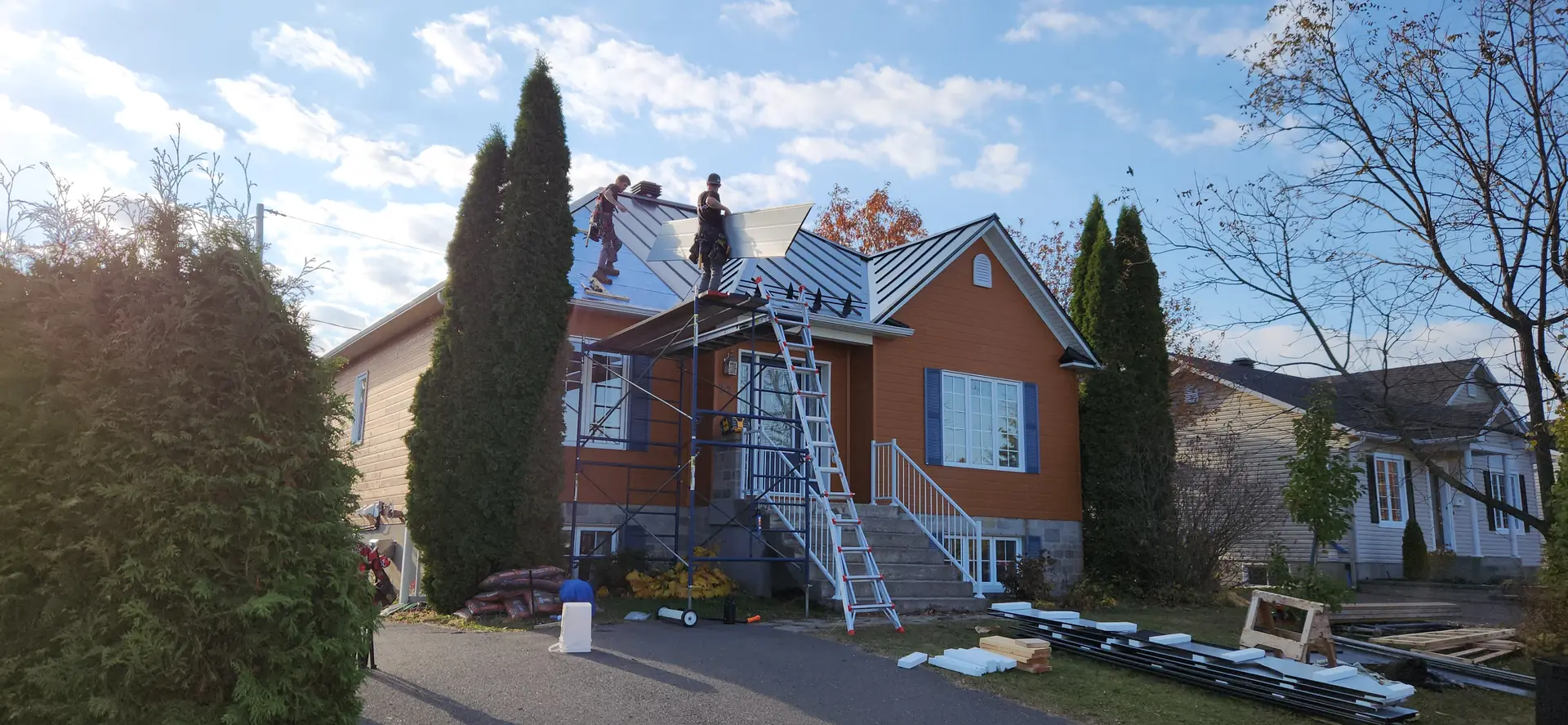 Roof renovation work on a residential house with workers installing metal panels, scaffolding, and equipment on site.