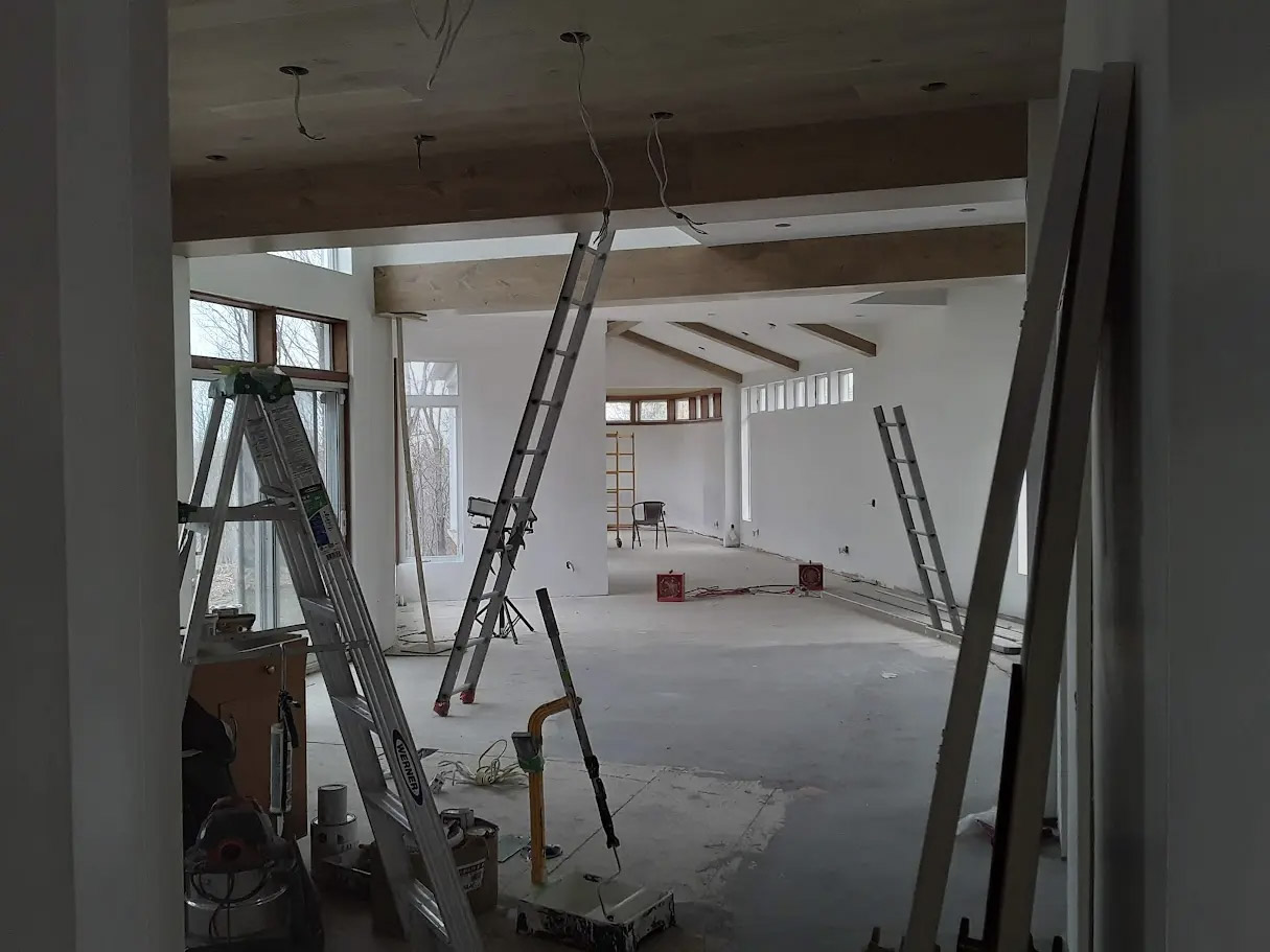 Home interior under renovation with high ceilings, exposed beams, ladders, hanging wiring, large windows, and drywall walls.