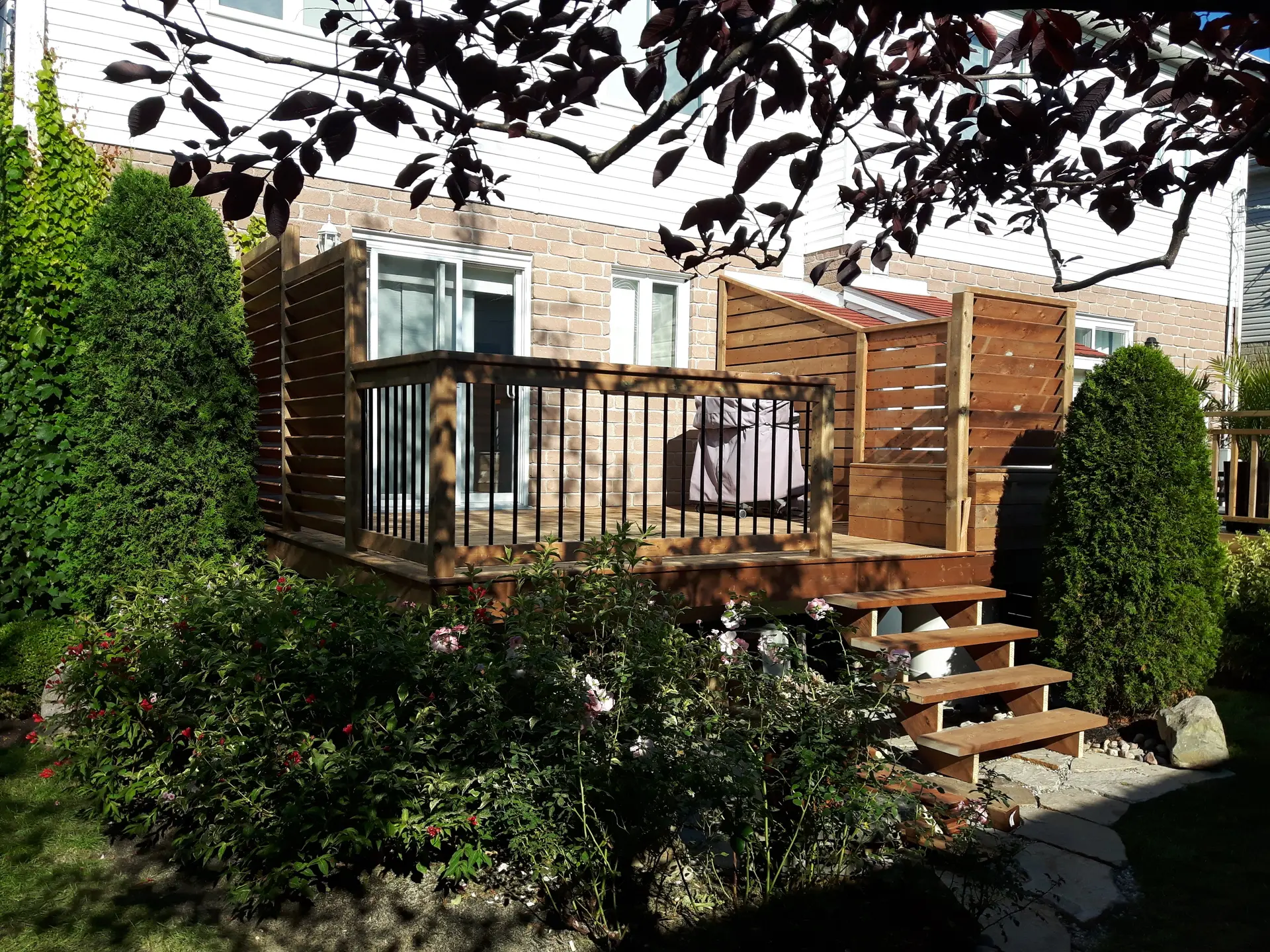 Wooden deck with metal railing, wooden slat privacy screen, and flower garden in front of a brick house.