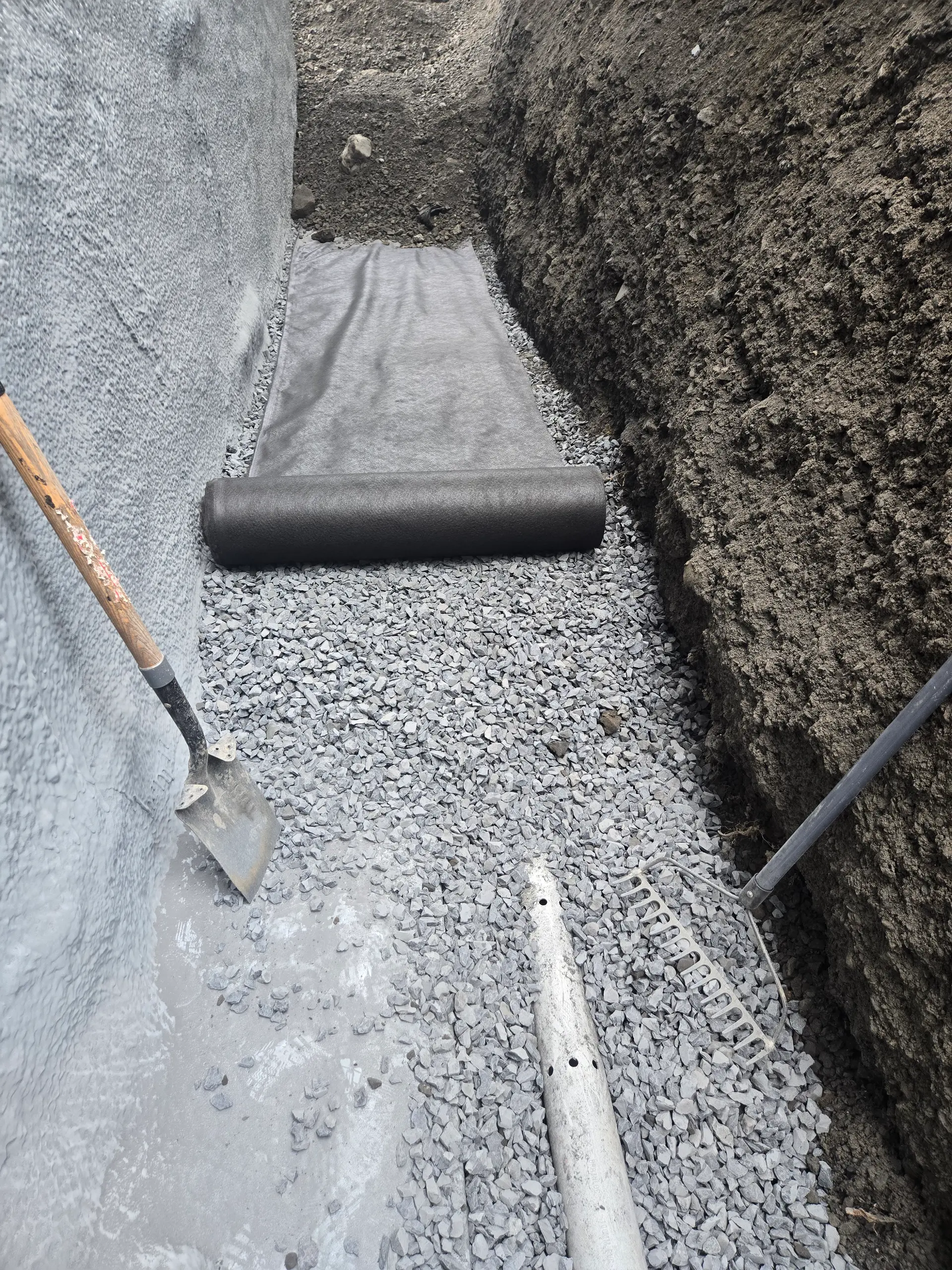 Excavation de fondation avec membrane géotextile, pierre concassée, tuyau de drain français perforé et outils de chantier pour drainage résidentiel