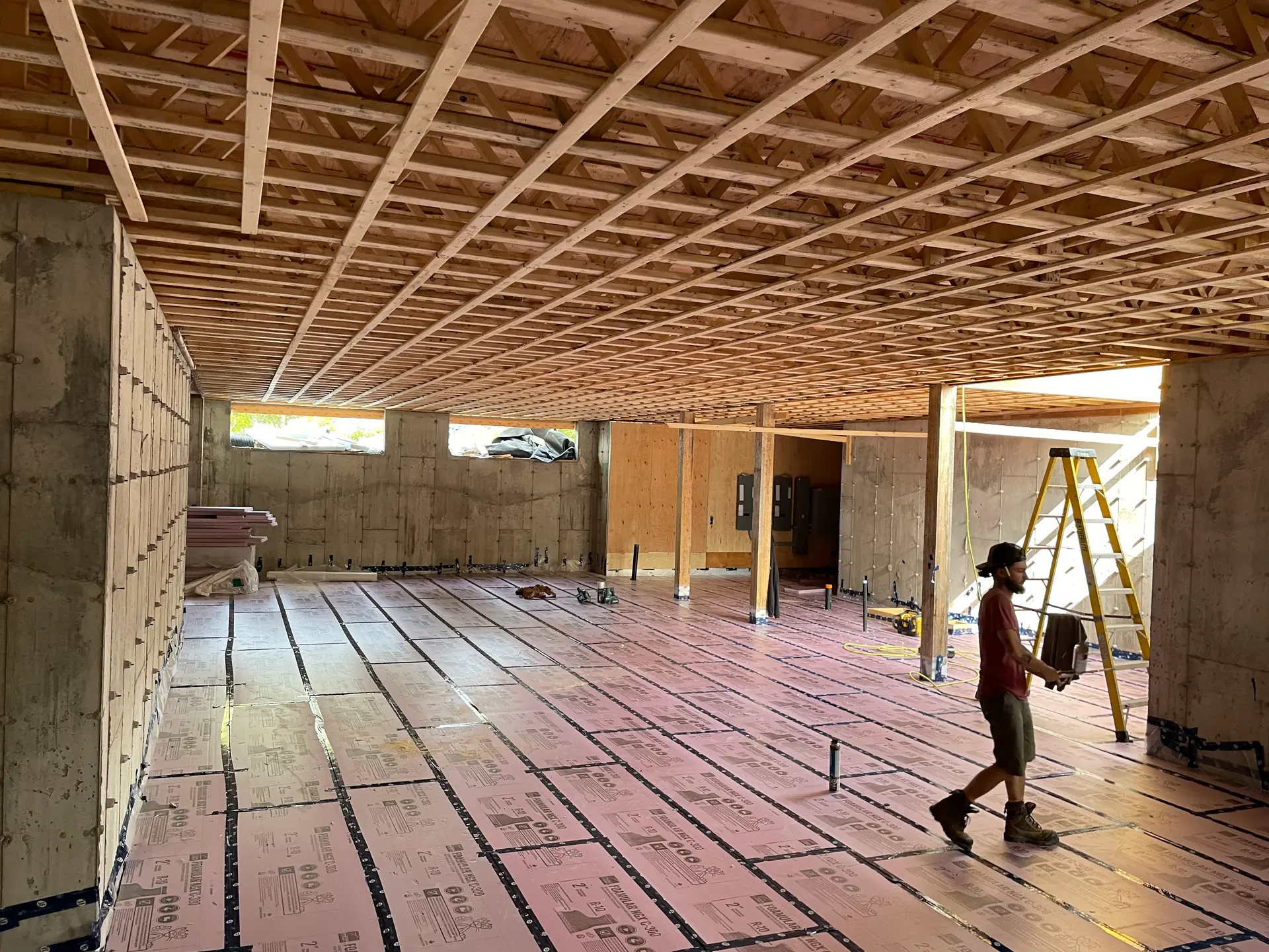 Sous-sol en construction avec isolation rigide au sol, charpente de plafond, murs de fondation et ouvrier sur chantier