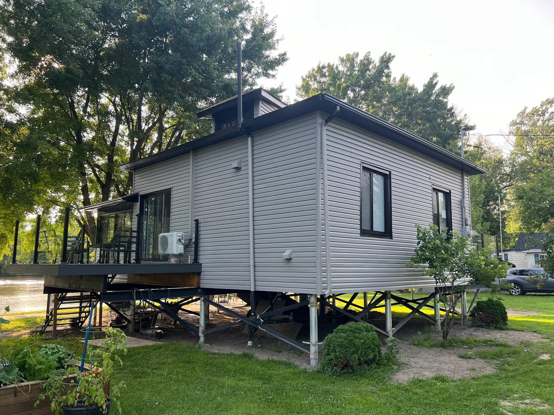 Maison sur pilotis avec revêtement extérieur gris, terrasse surélevée et structure métallique visible en milieu naturel