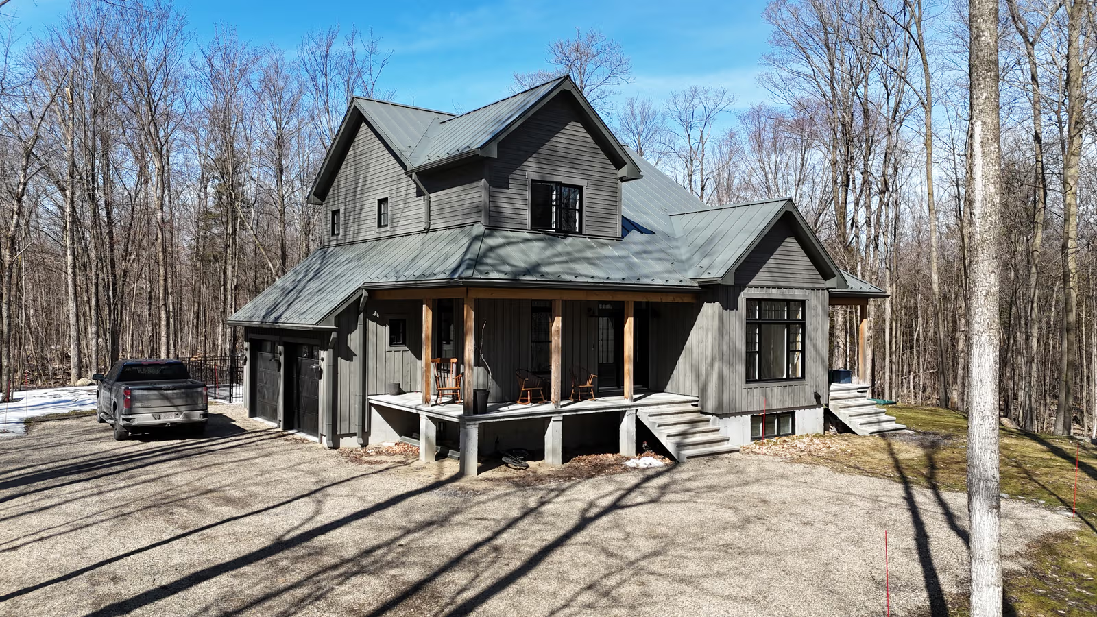 orest home with a grey metal roof, dark exterior siding, covered porch, attached garage, and gravel driveway.