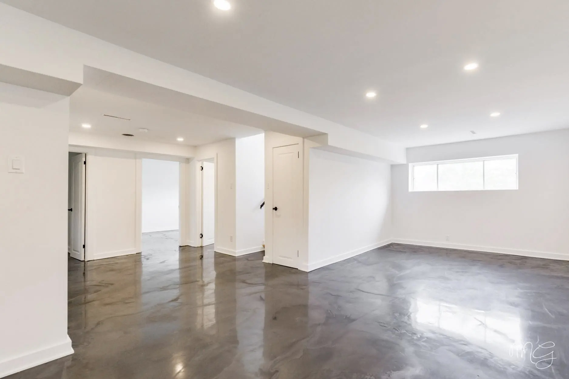 Sous-sol rénové avec plancher époxy gris lustré, murs blancs, portes intérieures, fenêtre horizontale et luminaires encastrés.