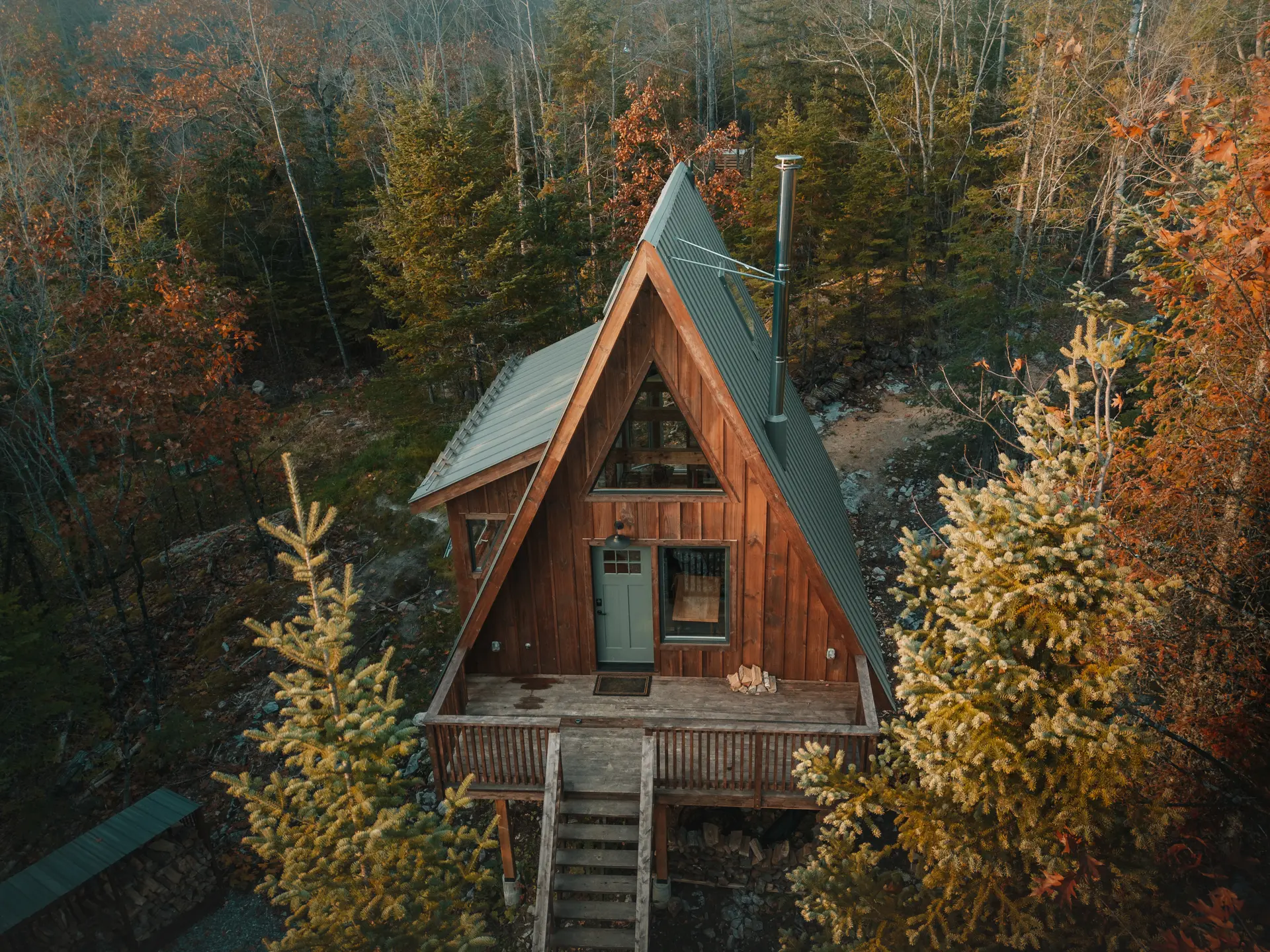 A-frame style wooden cabin nestled in an autumn forest with a metal roof and a wooden balcony.