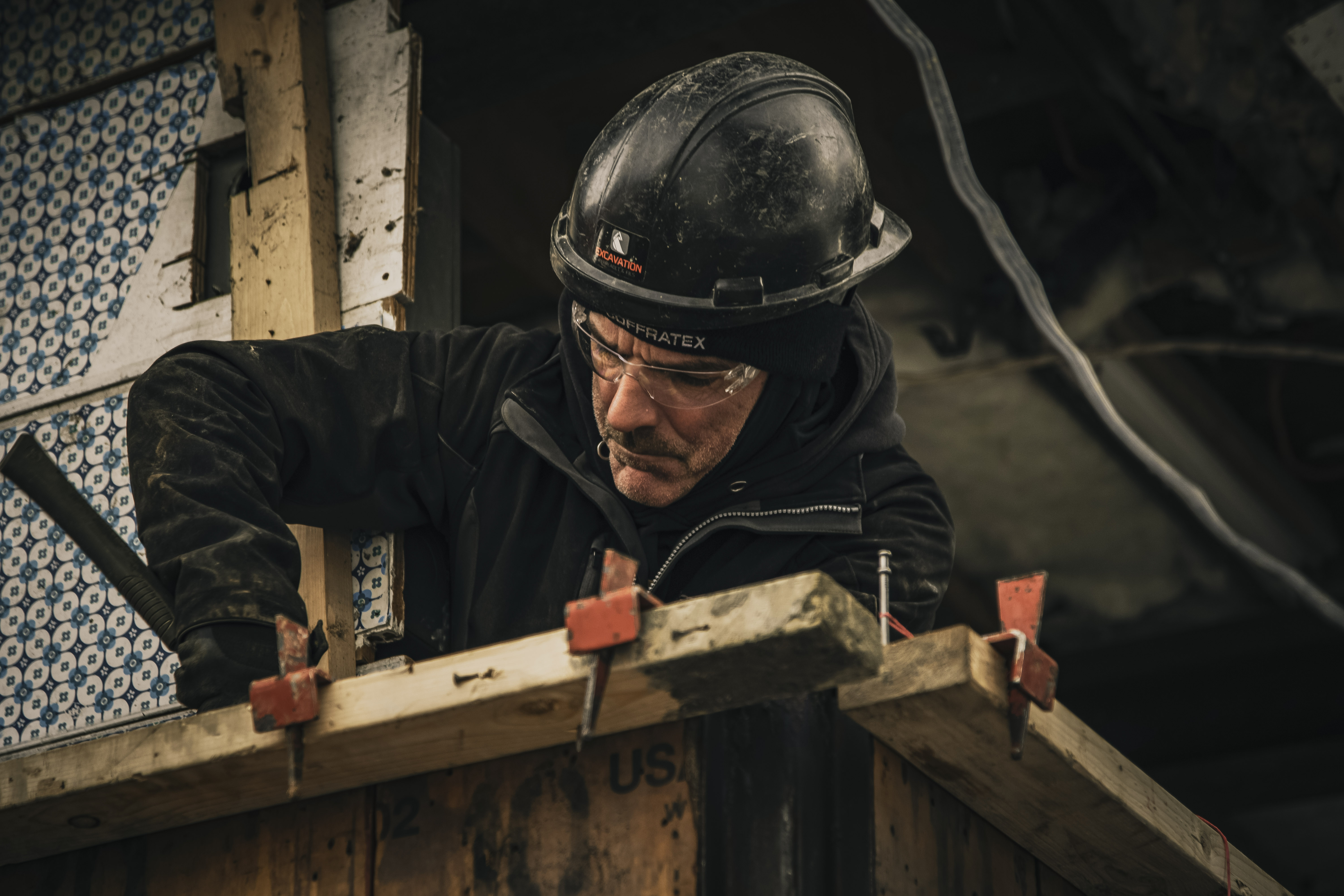 Worker wearing a black helmet and safety glasses, performing carpentry and construction work on a construction site.
