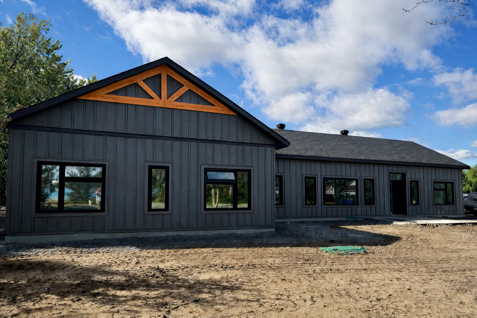 New contemporary home under construction with dark gray vertical siding, exposed wood trusses, shingle roof, large black-framed windows, and a graded construction site