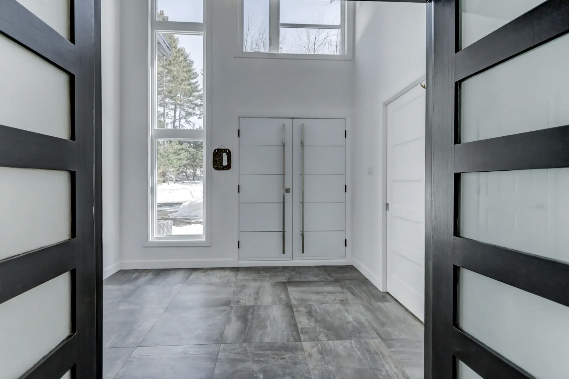 Bright modern entryway with white double doors, long metal handles, tall vertical windows, and gray tiled flooring.