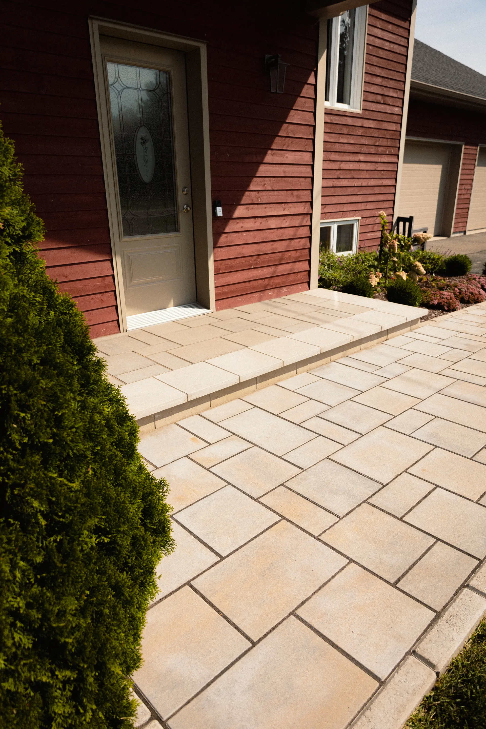 House entrance with beige interlocking pavers, slab step, glass front door, red siding, and landscaping.