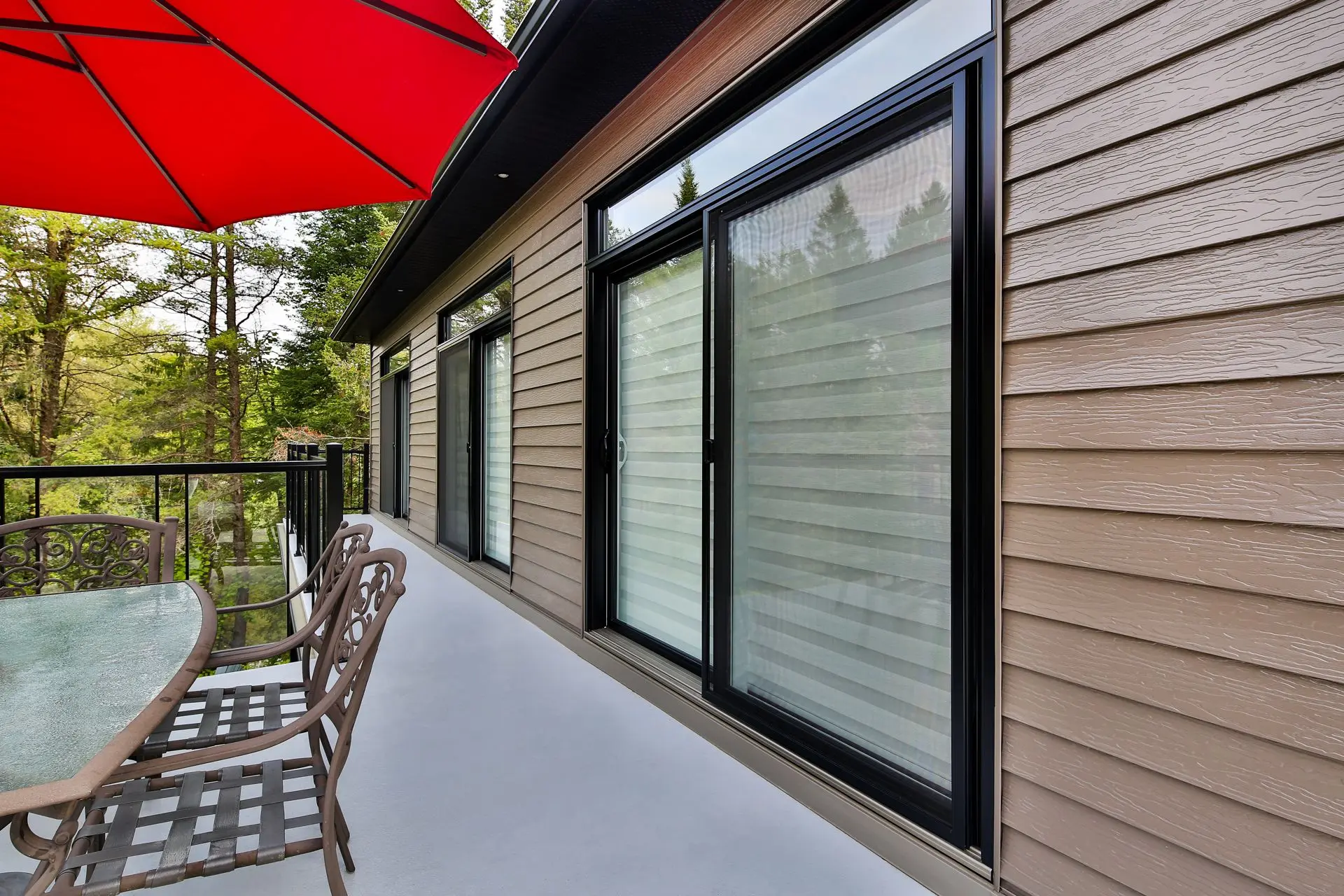Terrasse extérieure moderne avec revêtement en bois, grandes fenêtres coulissantes et mobilier de terrasse en métal sous un parasol rouge.