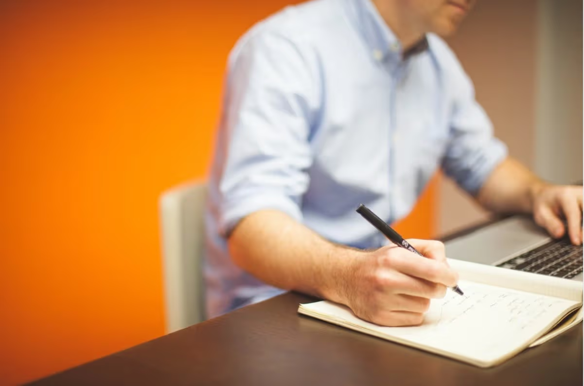 Professionnel prenant des notes dans un carnet à côté d’un ordinateur portable, dans un bureau moderne avec un mur orange.
